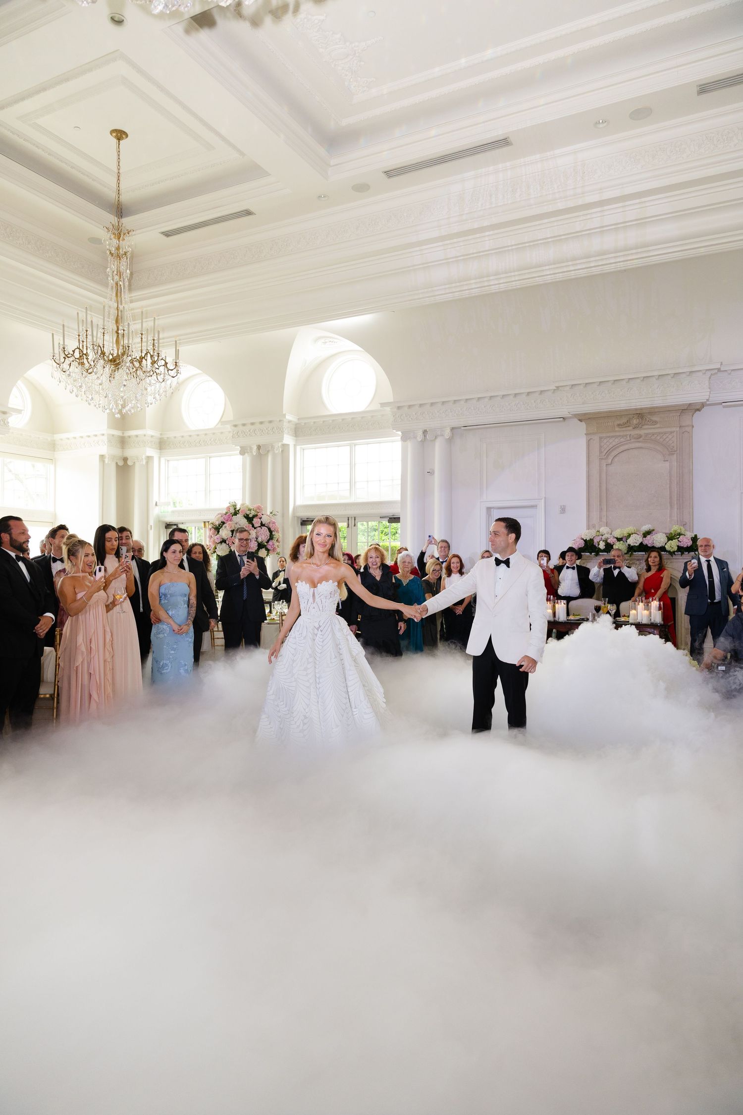 A couple shares their first dance in an elegant white ballroom with guests watching from the sides as fog covers the floor.