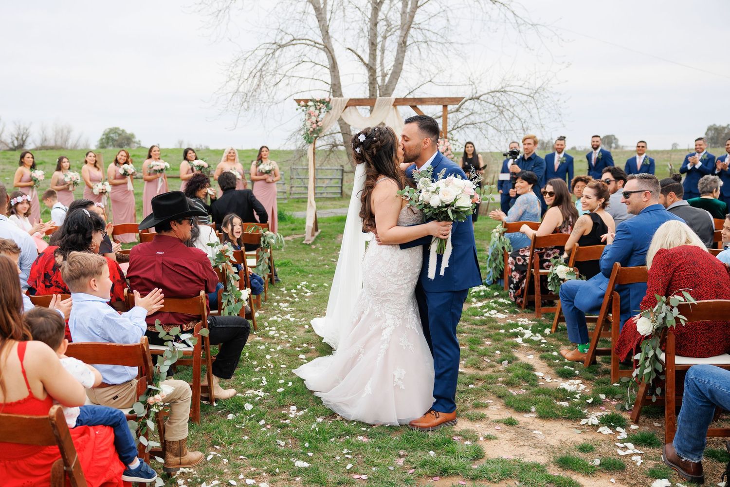 Guests sit in rows of wooden chairs at an outdoor wedding ceremony with scattered flower petals on the ground.