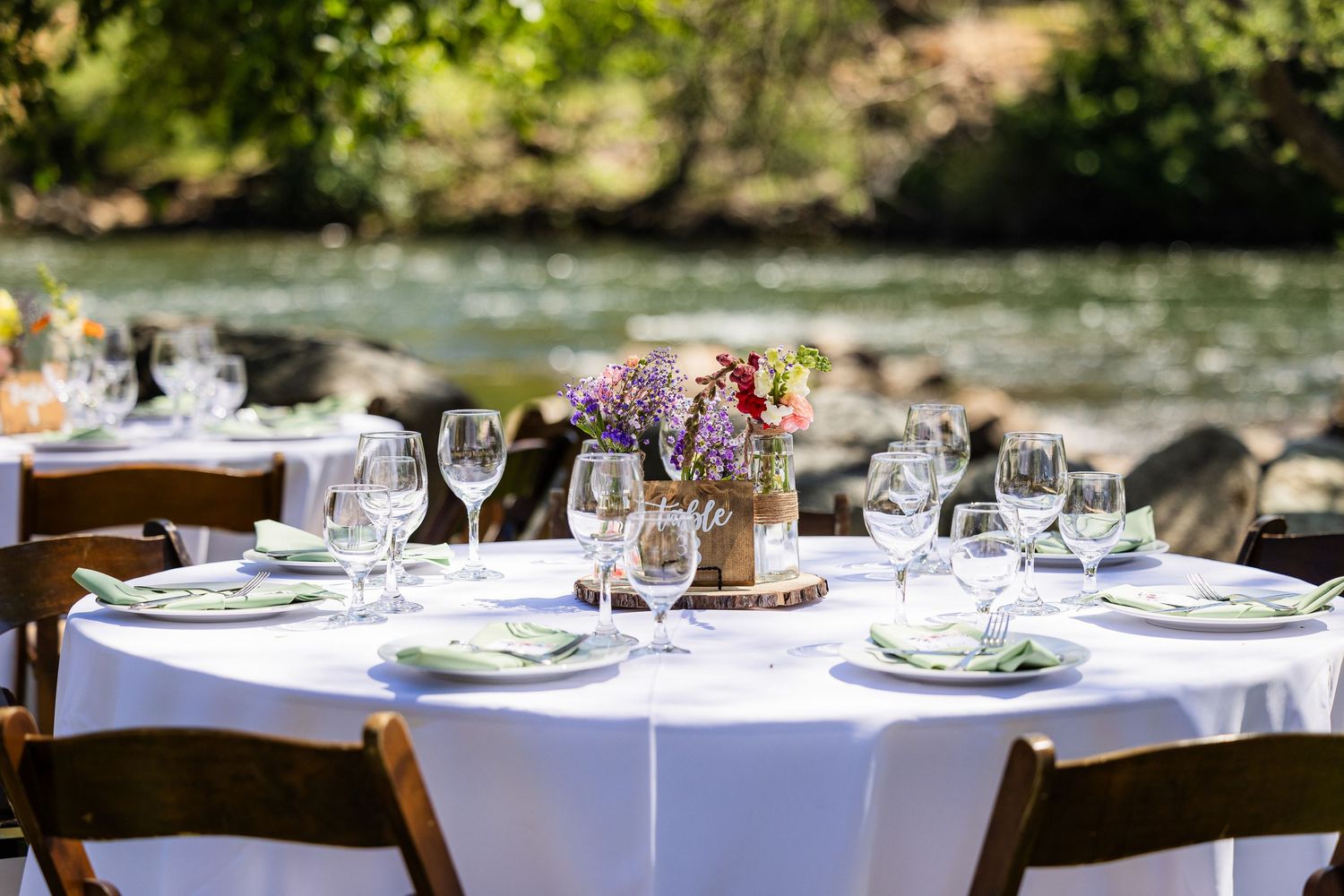 Elegant outdoor wedding reception tables with white linens, wine glasses, and floral centerpieces in a garden setting.