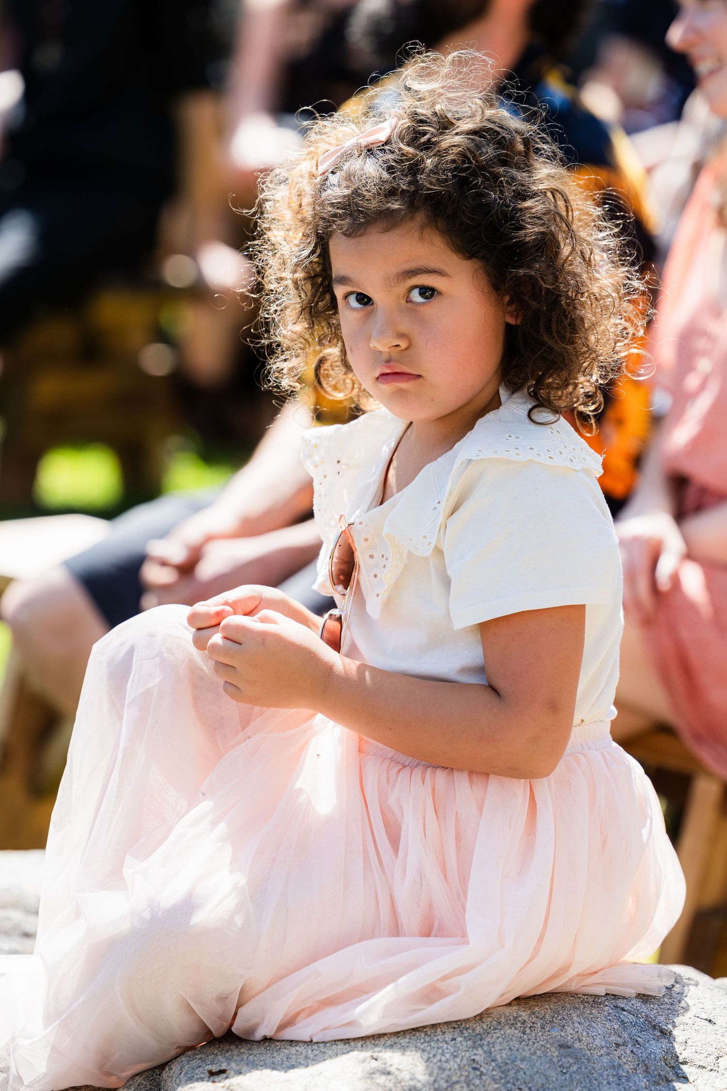 A young child in a white and pink dress sits gracefully on a stone surface in a sunlit outdoor setting.
