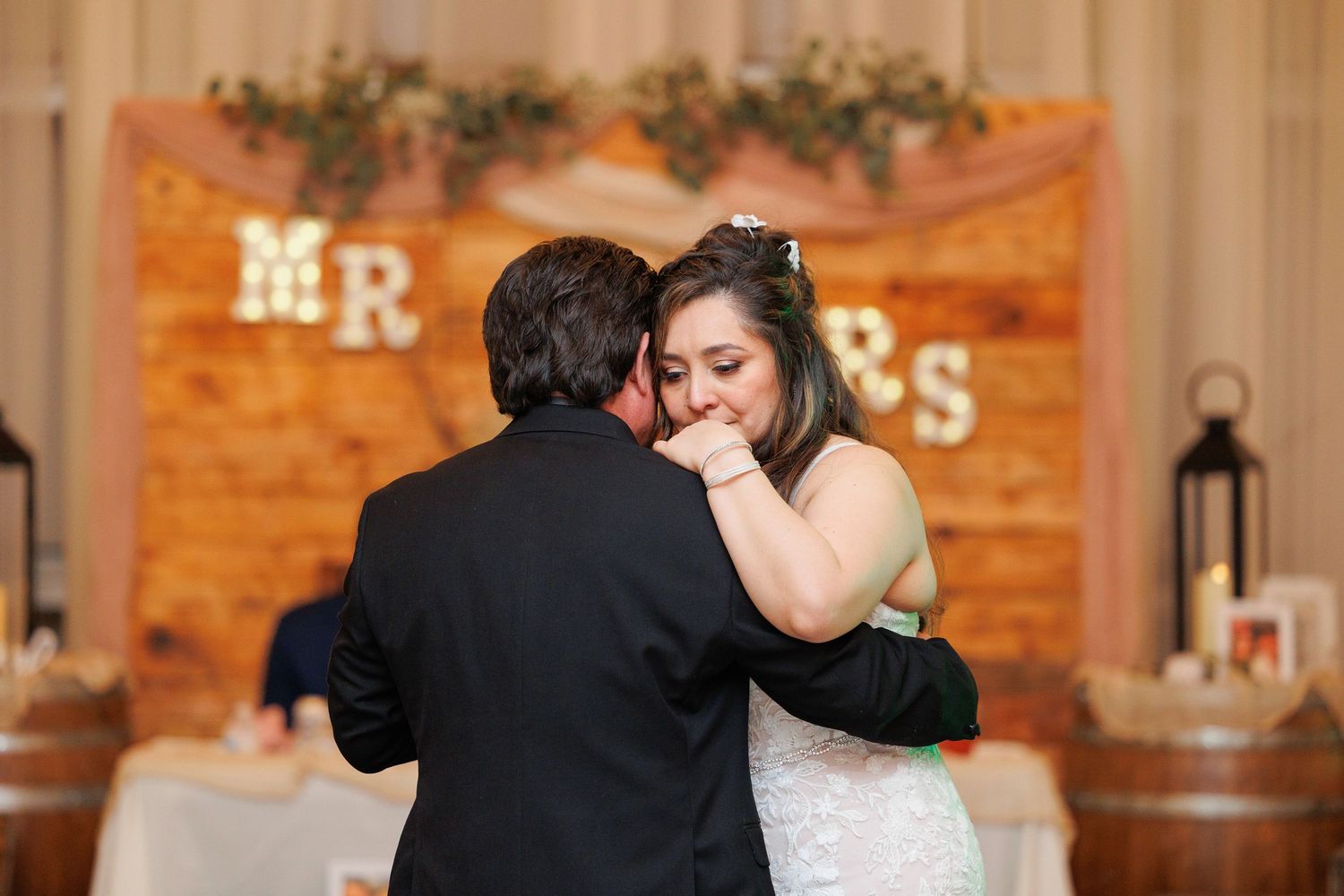 A couple shares their first dance at a wedding reception with rustic wood wall decor and string lights in the background.
