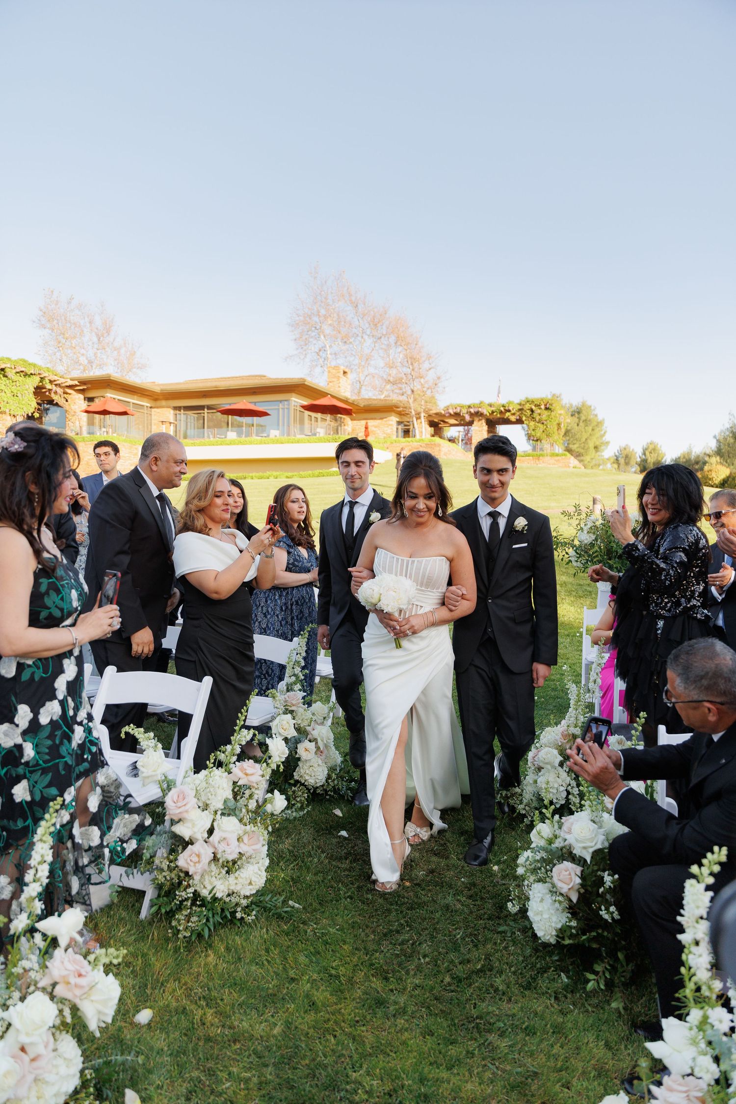 Wedding guests toss confetti and celebrate as newlyweds walk through floral arch during outdoor ceremony.