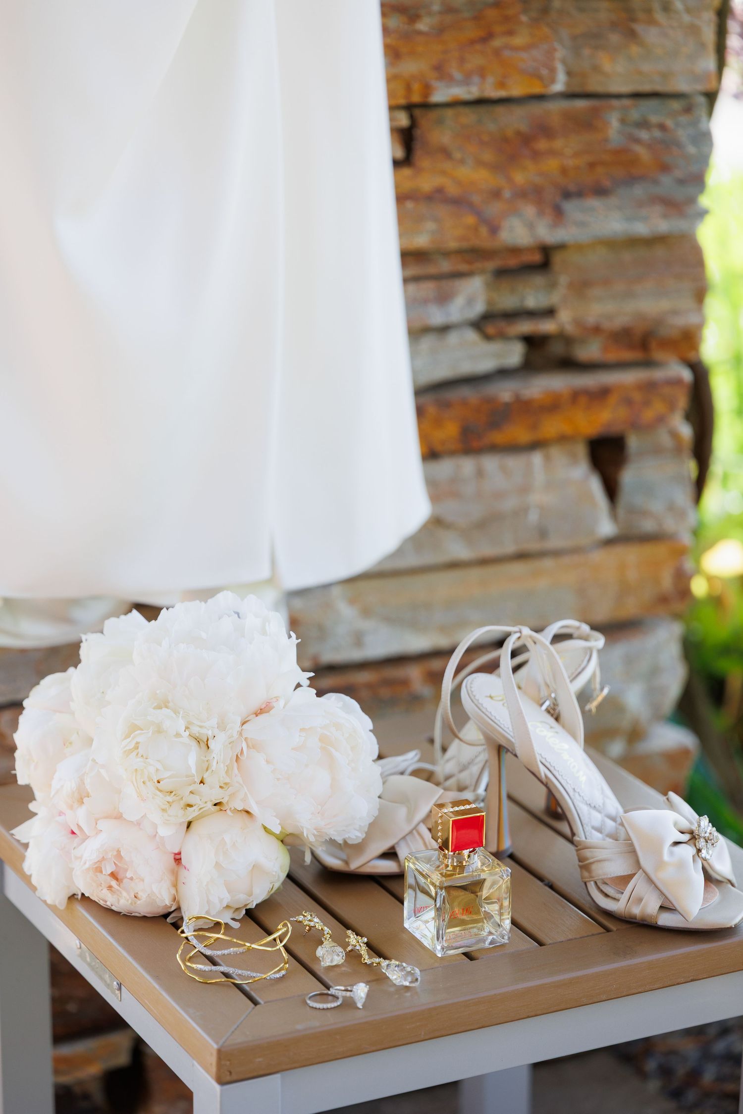 Wedding accessories including white floral bouquet, perfume bottle and bridal sandals arranged on wooden table with stone wall.