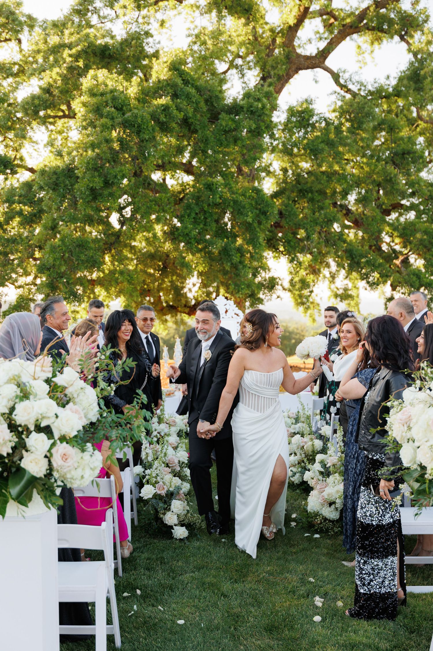 Wedding guests mingle and celebrate outdoors under a large tree with elegant white floral arrangements and chairs.