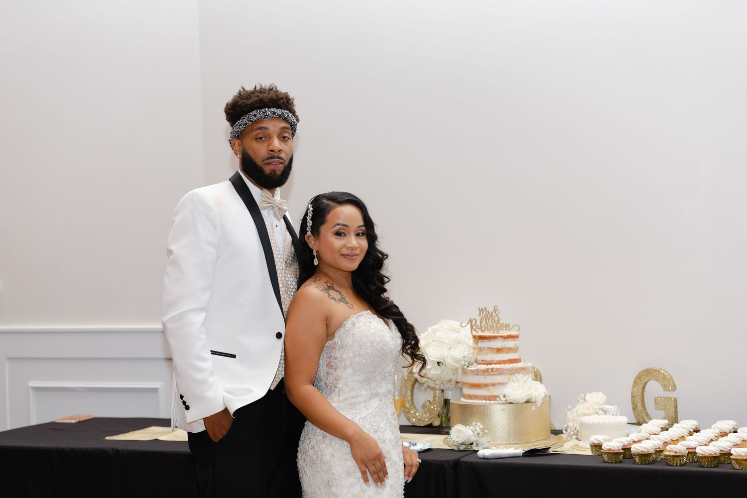 A couple poses next to their elegant naked wedding cake display with gold decorative elements on a black table.