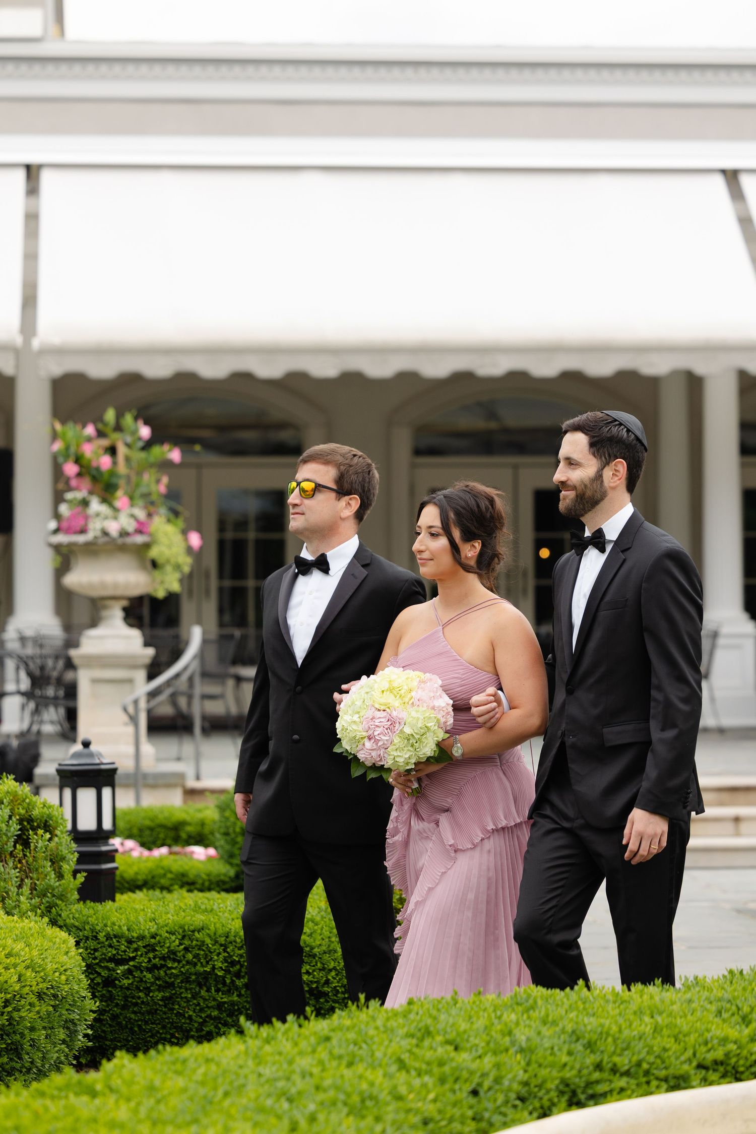 Wedding party in formal attire poses outdoors with bridesmaids in pink dresses and groomsmen in black tuxedos.