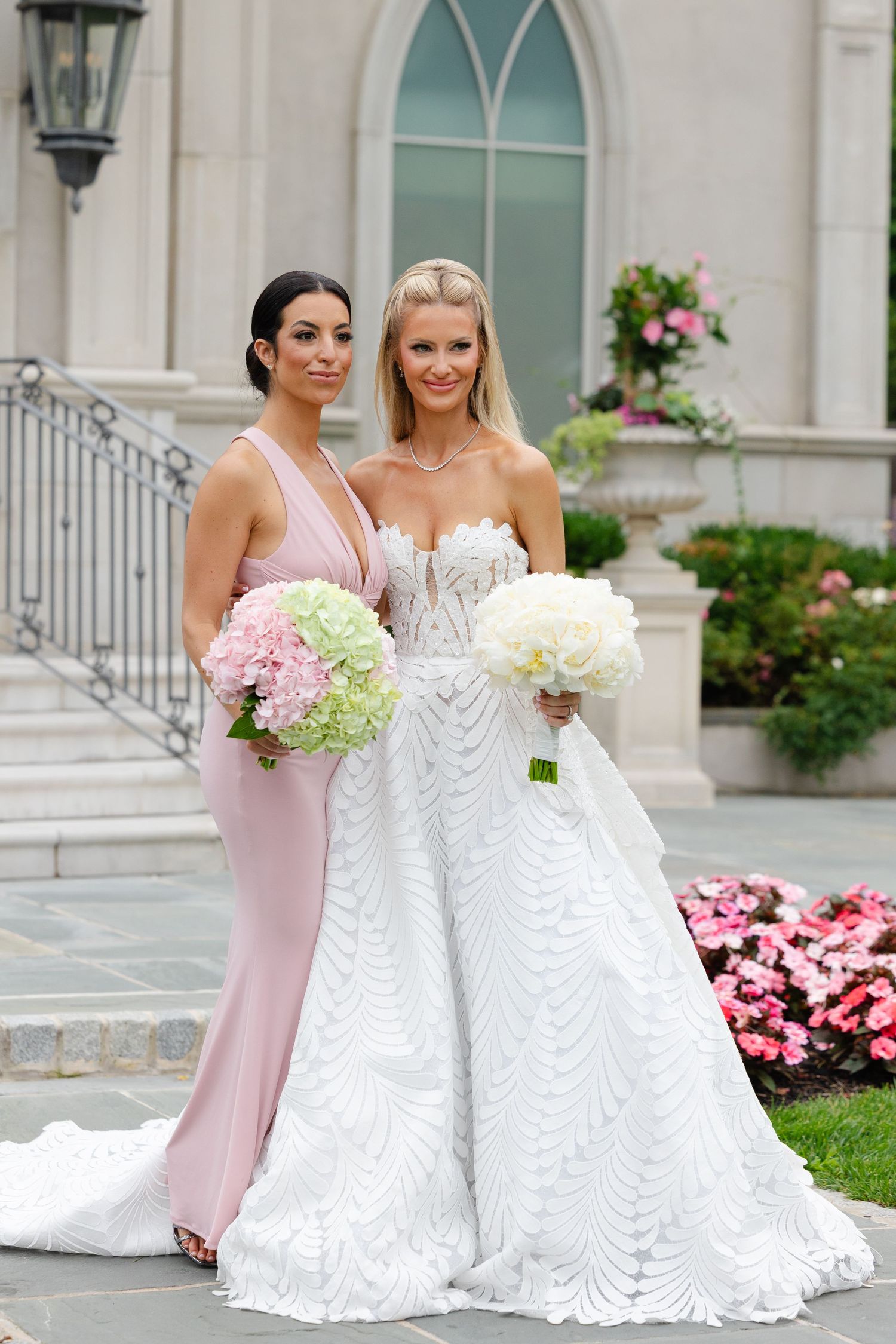 Wedding party members in blush pink and white gowns pose together at an elegant venue with blooming flowers.