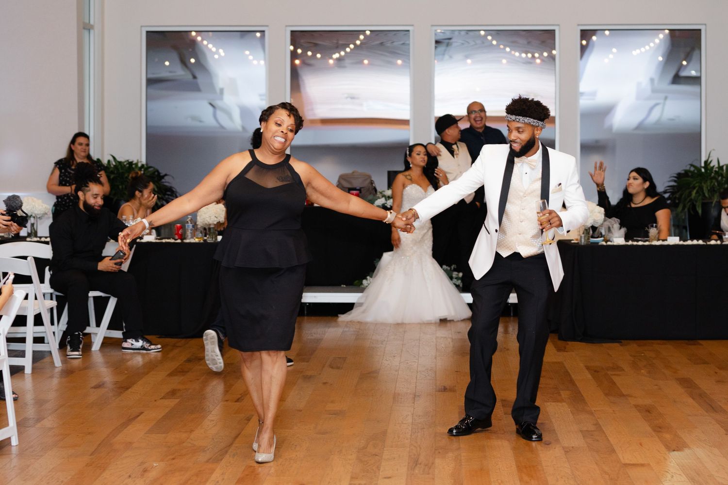 Wedding guests perform a coordinated dance routine in a reception hall with string lights and wooden floors.