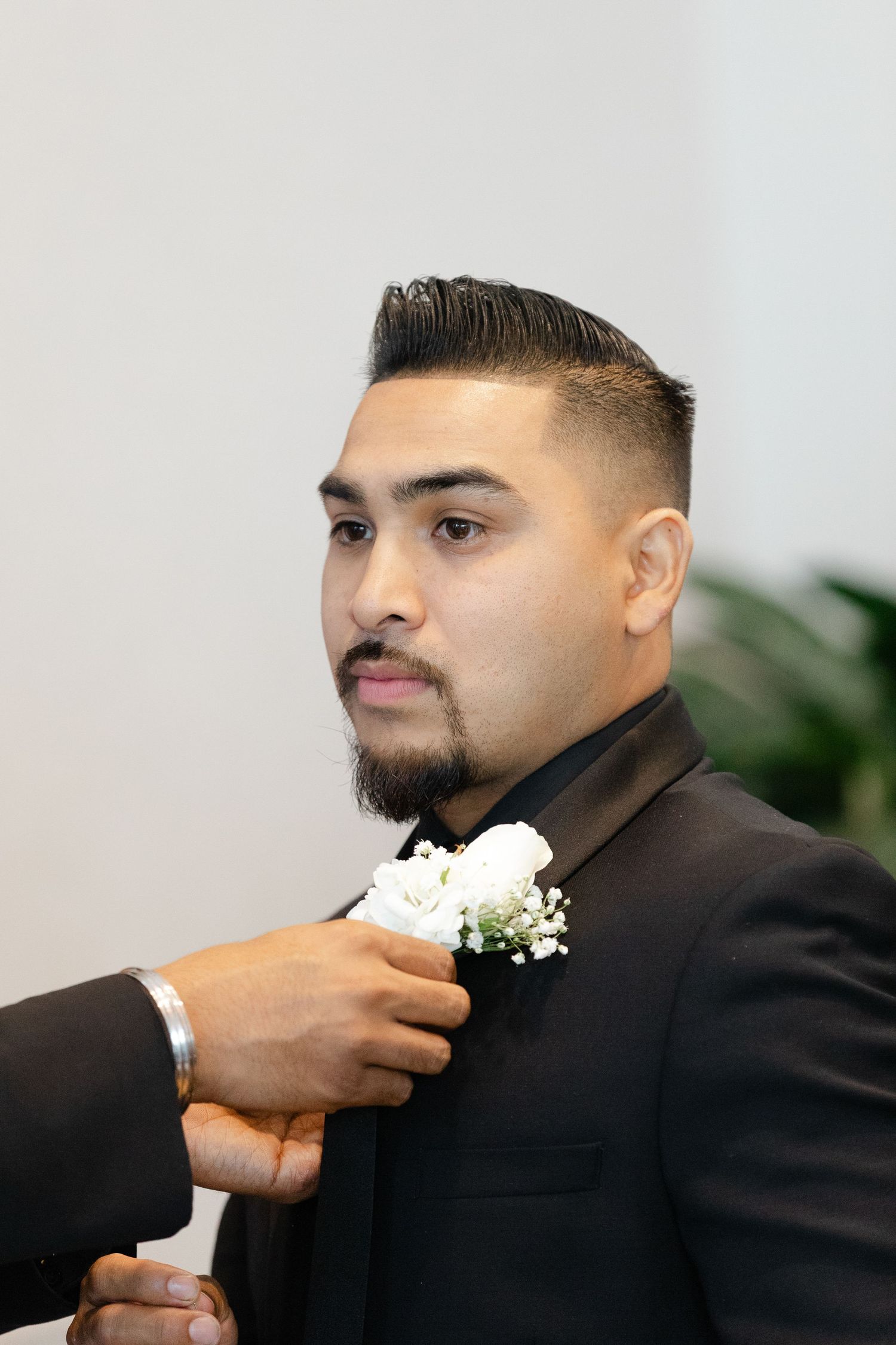 Someone helps adjust a white boutonniere flower on a black tuxedo jacket at a formal event.