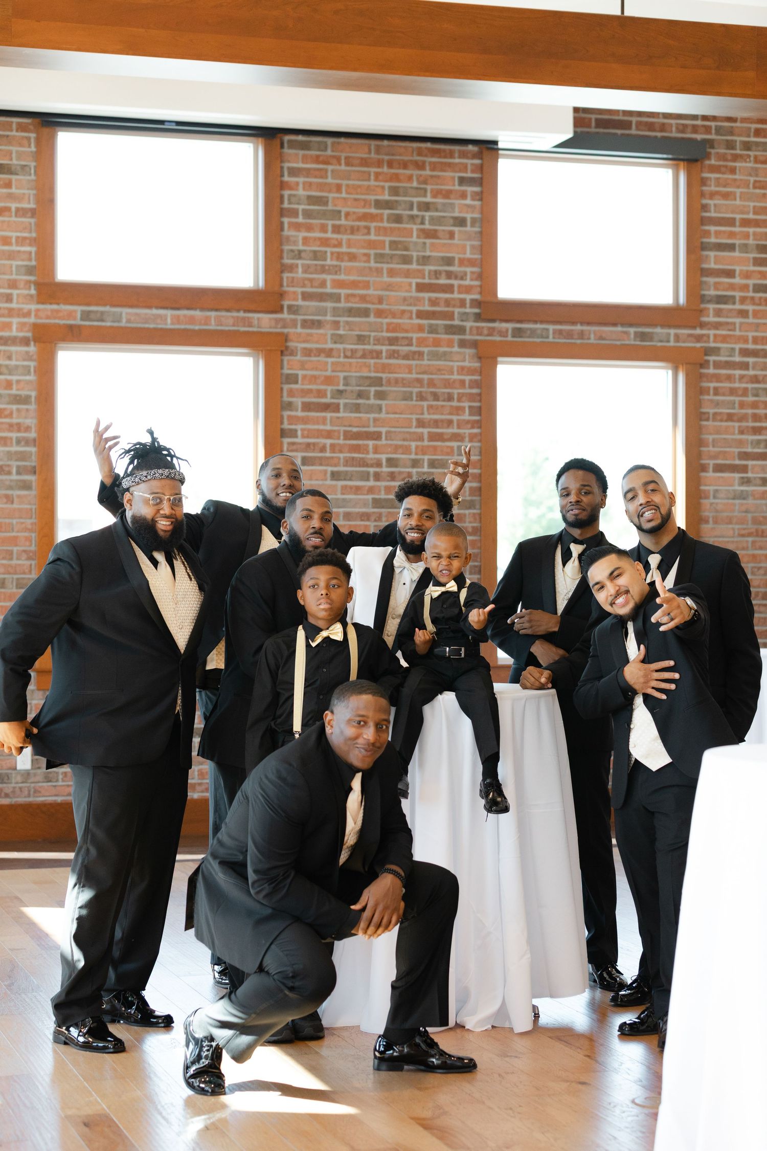 A group of groomsmen in black tuxedos strike playful poses during a wedding photo session in a brick-walled venue.