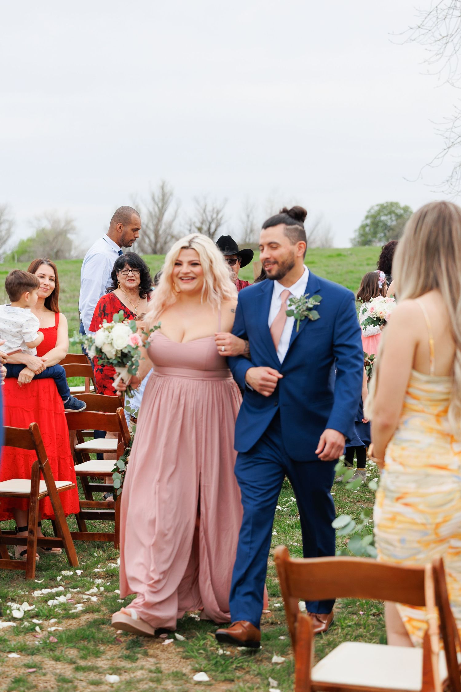 Wedding guests dance and celebrate at an outdoor reception with wooden chairs and scattered flower petals on the ground.