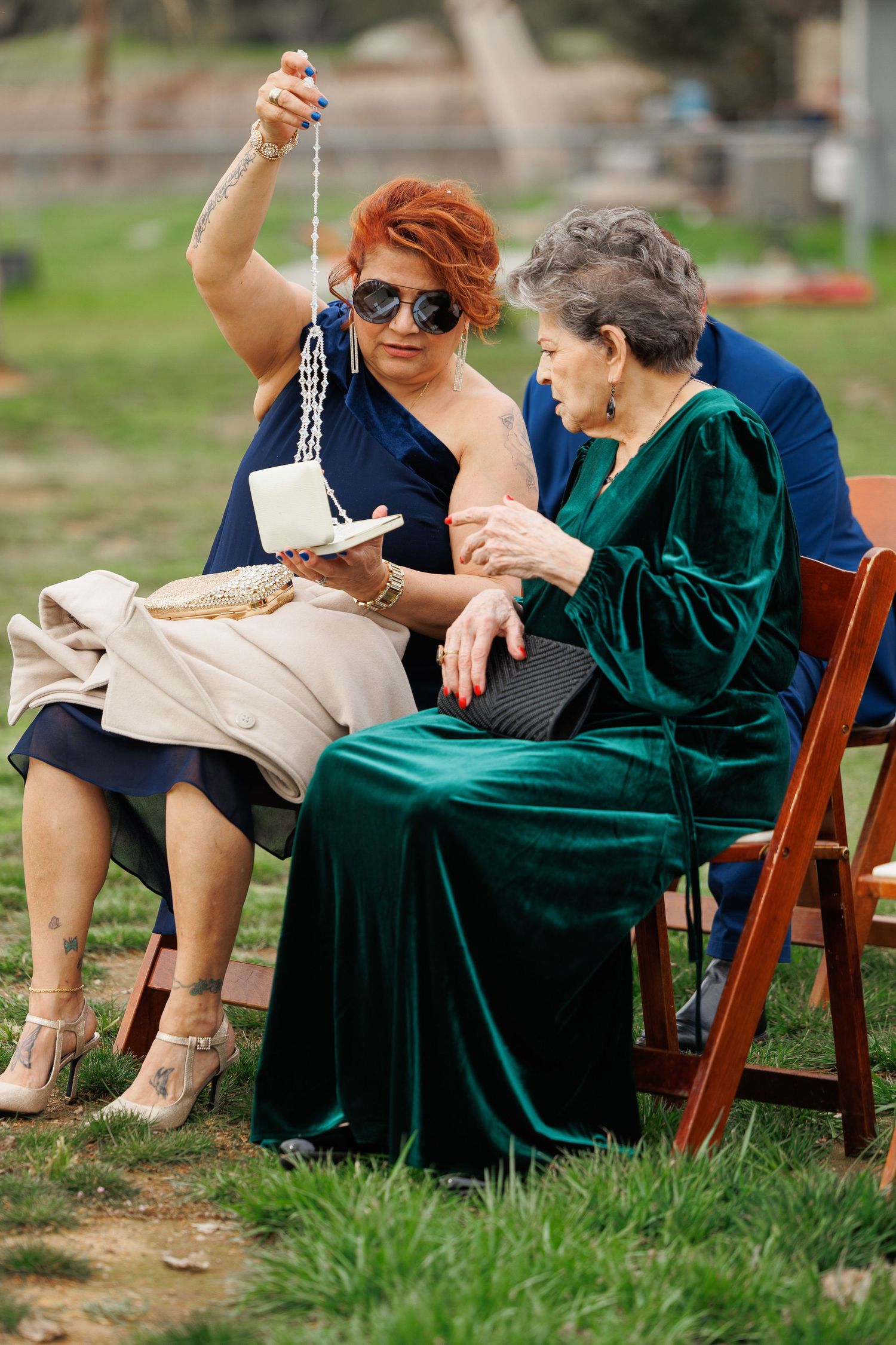 Two people in casual and formal attire sit together on wooden chairs in a grassy outdoor setting.