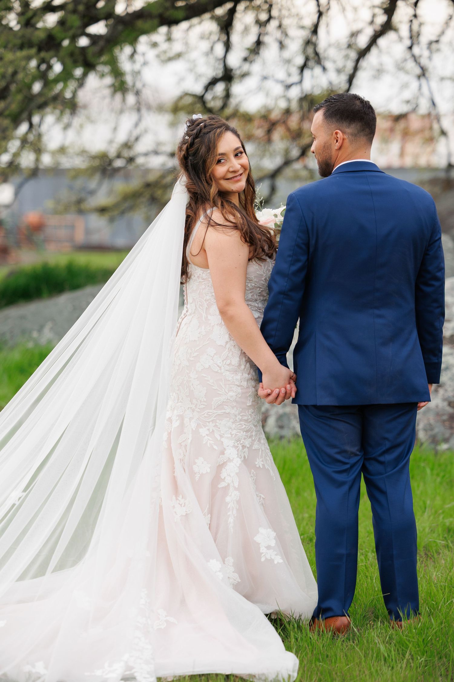 Newlyweds in navy suit and white lace wedding dress pose together on grassy hillside with flowing veil in spring setting.