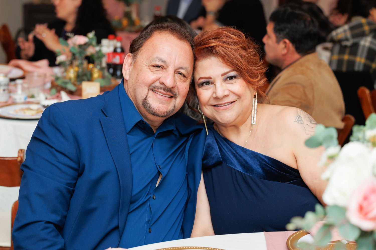 A couple in blue attire share joyful moments at a wedding reception with floral decor in the background.