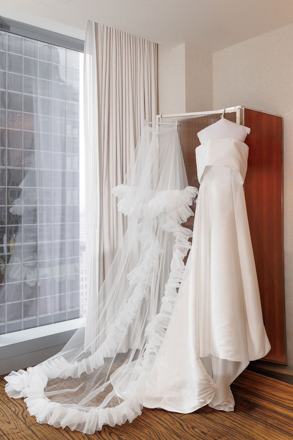 Elegant white wedding dress and flowing veil displayed on wooden hangers against light curtains.