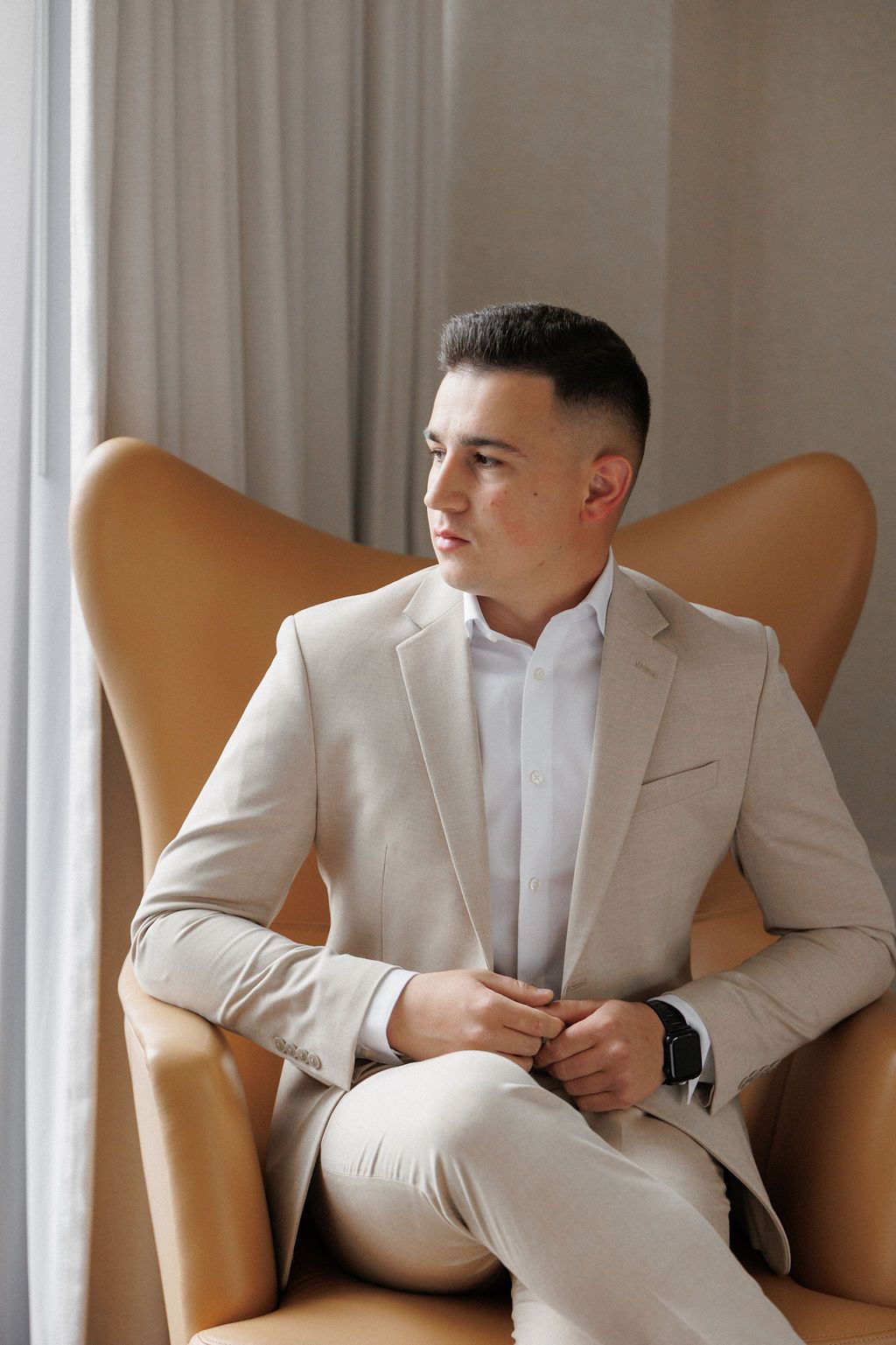A man in a light beige suit sits contemplatively in a tan leather chair by a window.