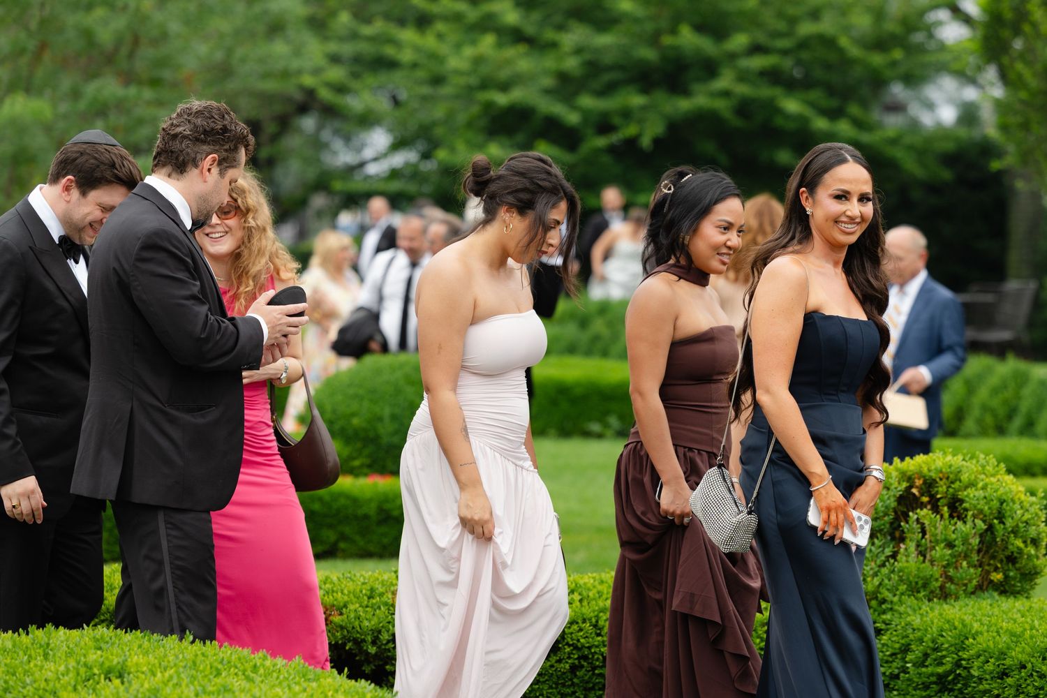 Elegantly dressed wedding guests in formal attire walk through lush green gardens at an outdoor reception.
