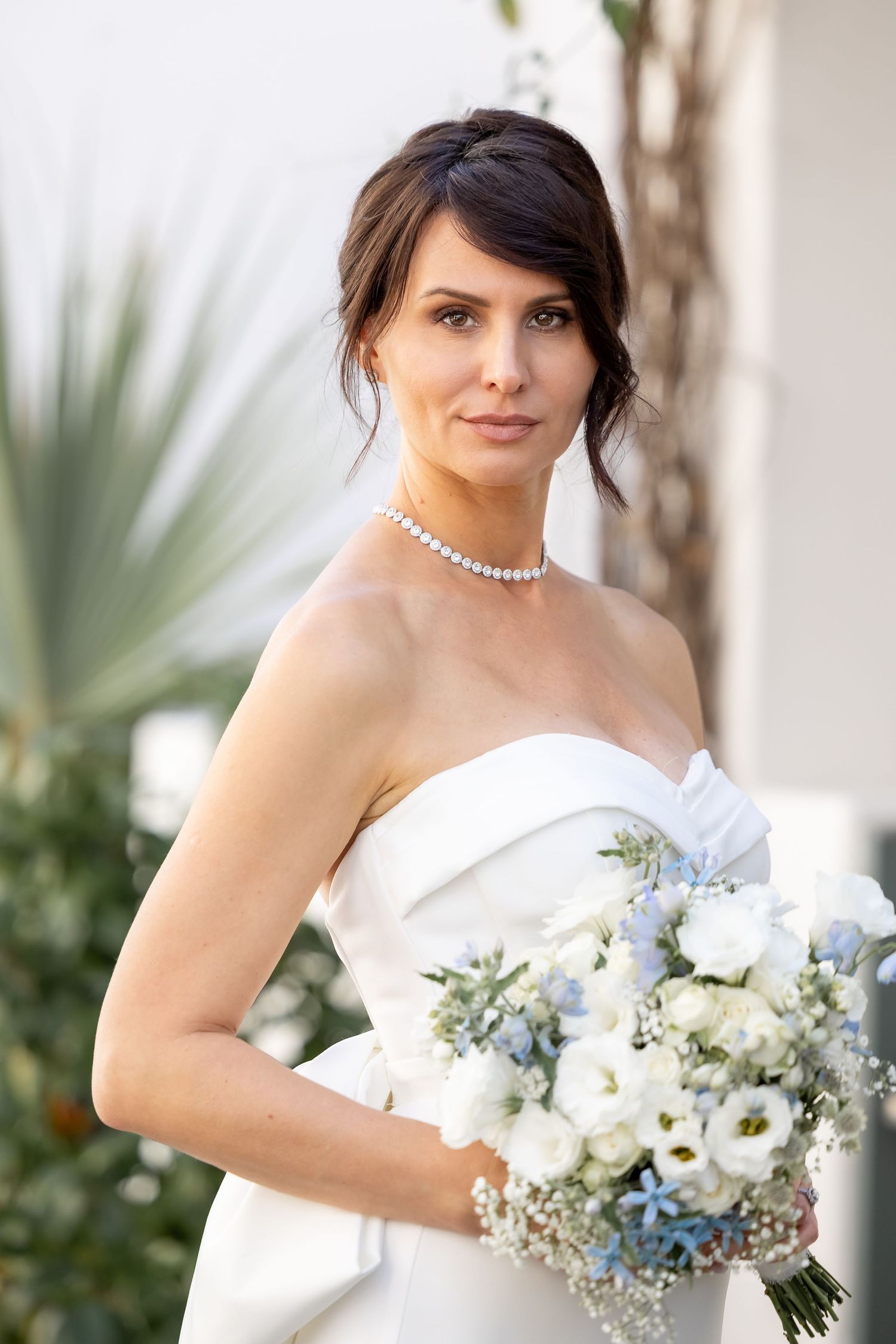 Close-up bridal portrait showcasing white and blue floral bouquet with pearl necklace and strapless gown.