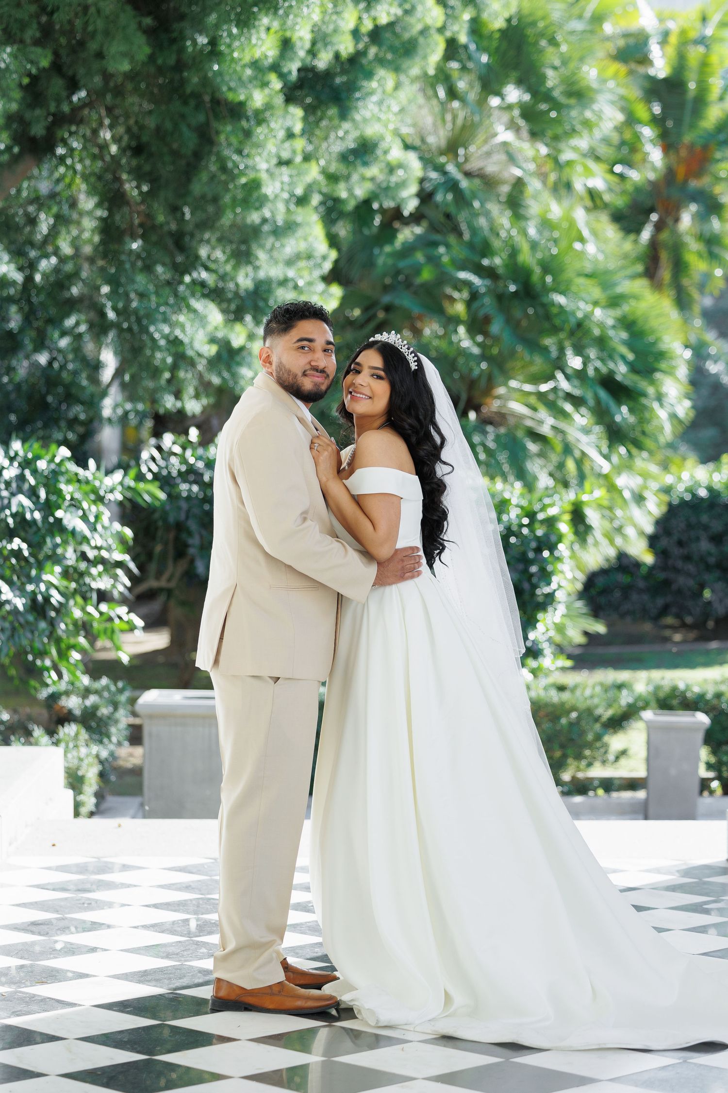 Wedding couple in white attire shares intimate moment on checkered outdoor patio.