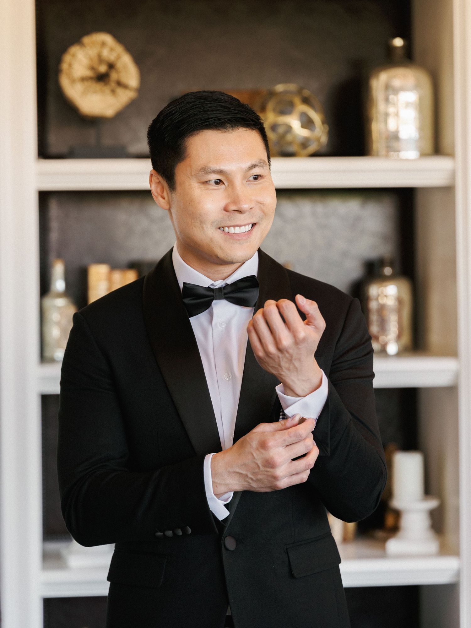Groom in classic black tuxedo and bow tie adjusts cufflink while smiling in front of decorative shelving.