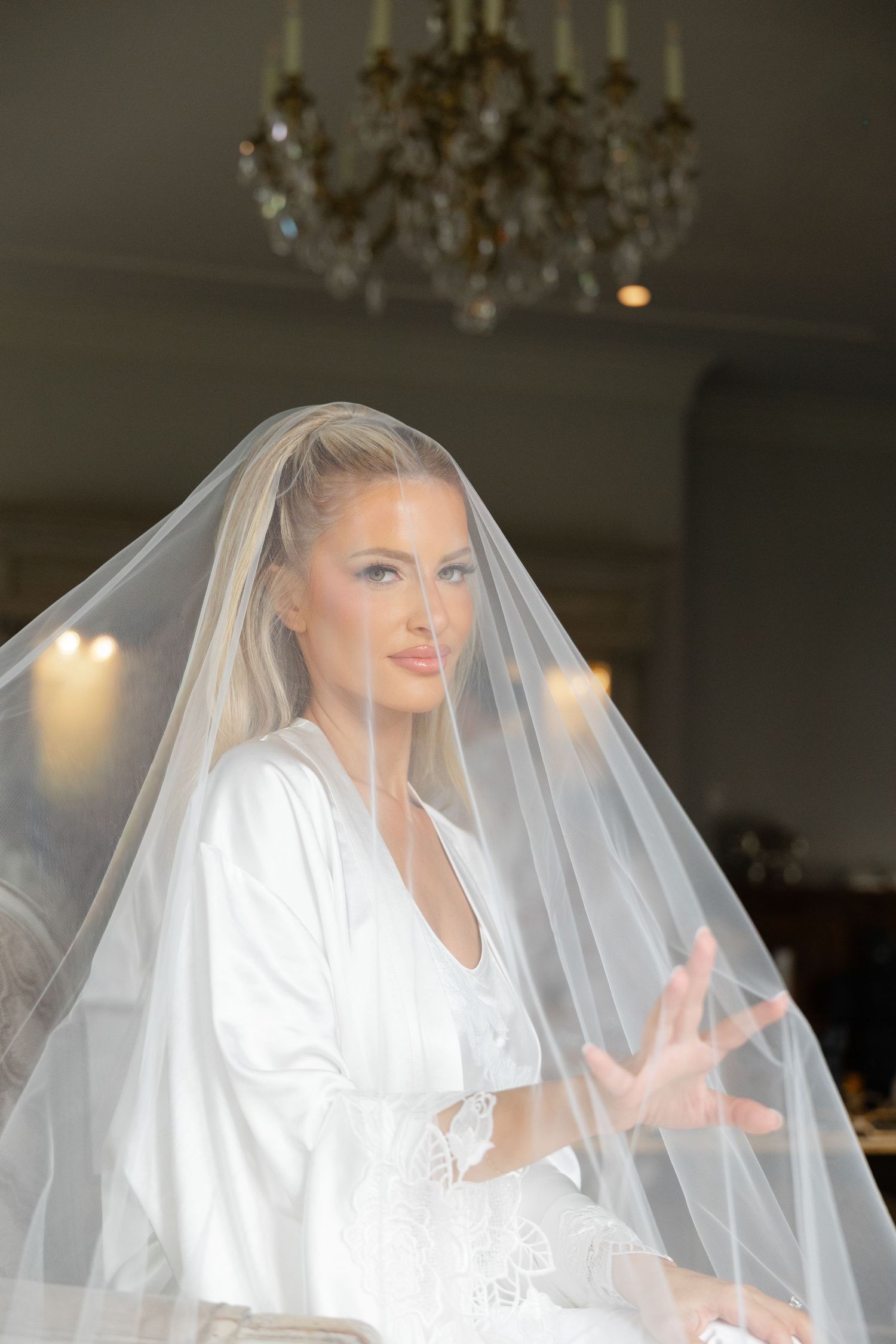 Bride in white silk robe and sheer veil poses elegantly under crystal chandelier during bridal preparation.