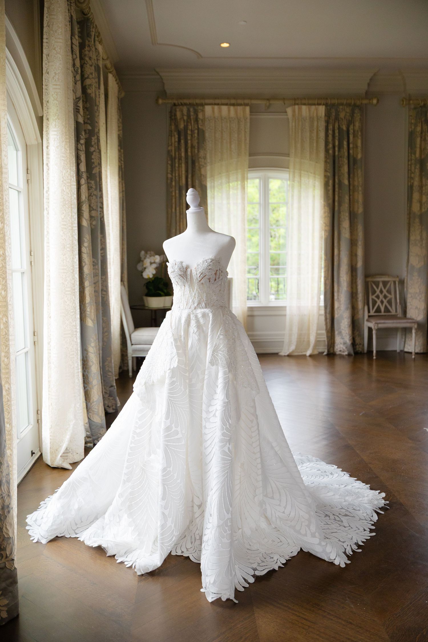 Elegant white lace wedding gown with full skirt and train displayed on mannequin in sunlit room with flowing curtains.