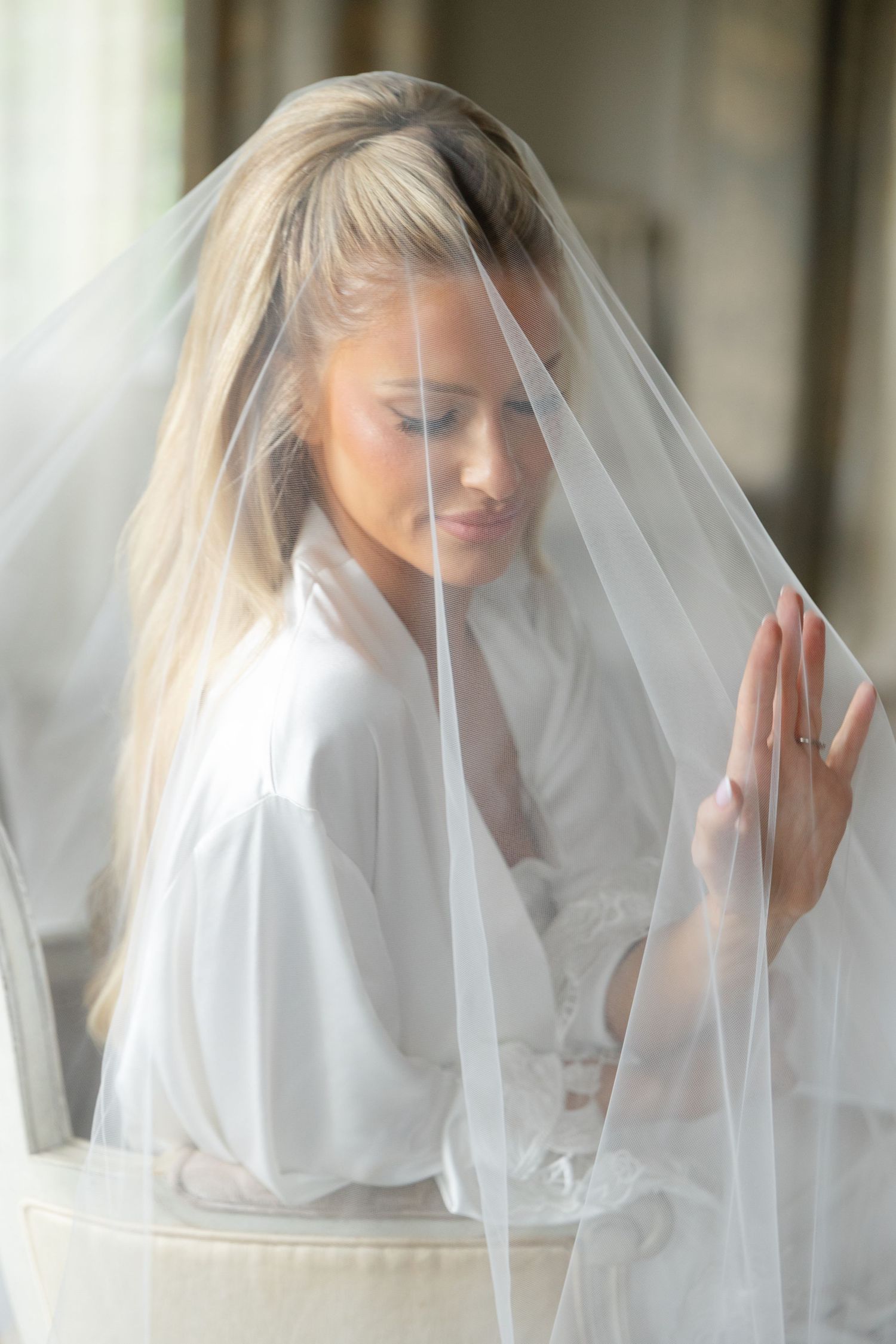 Elegant bridal portrait showing delicate white veil and robe in soft natural window light.