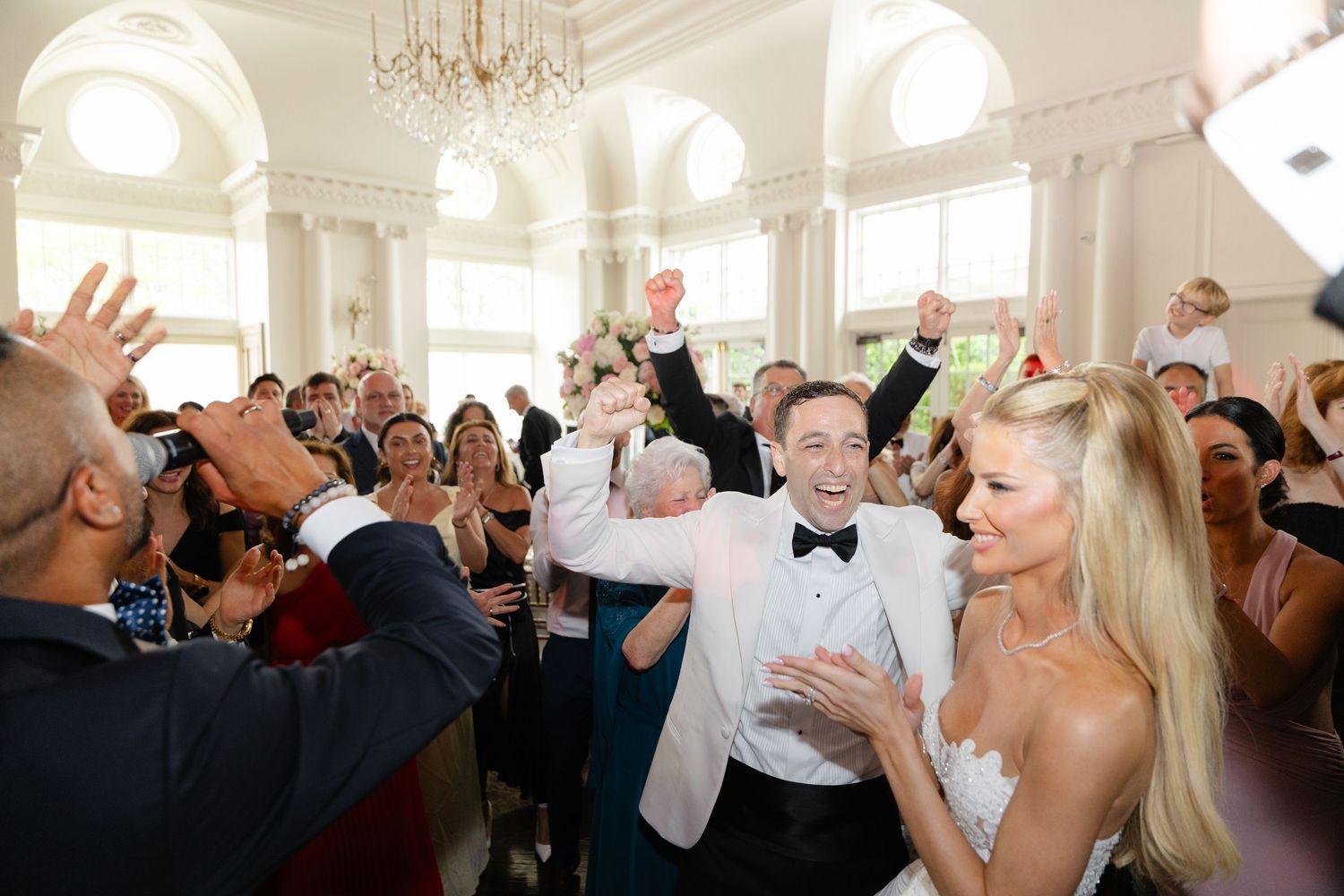 Wedding guests raise their glasses in celebration during an elegant reception in a bright white ballroom.