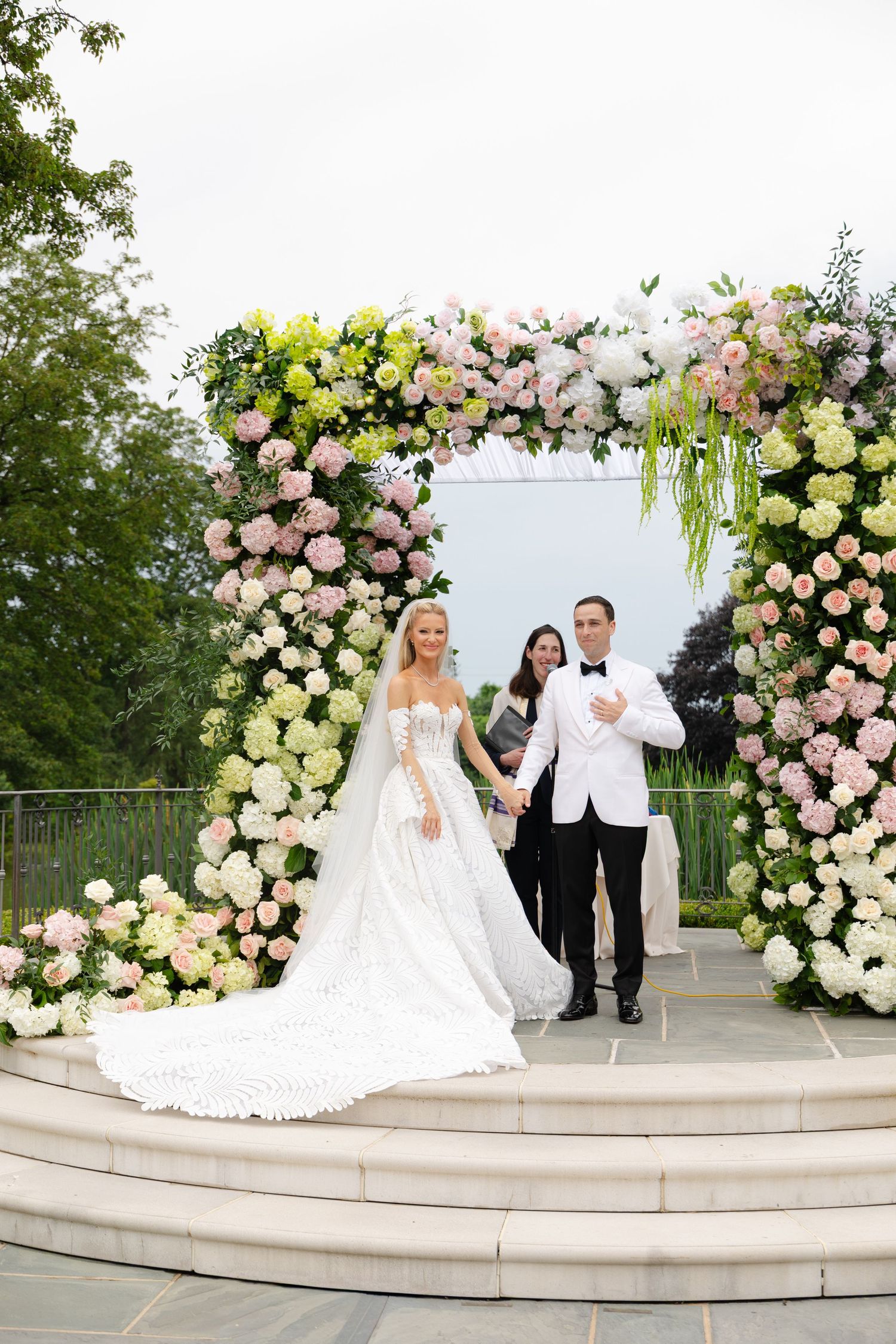Elegant wedding ceremony under a lush floral arch with pink and white roses on curved stone steps outdoors.
