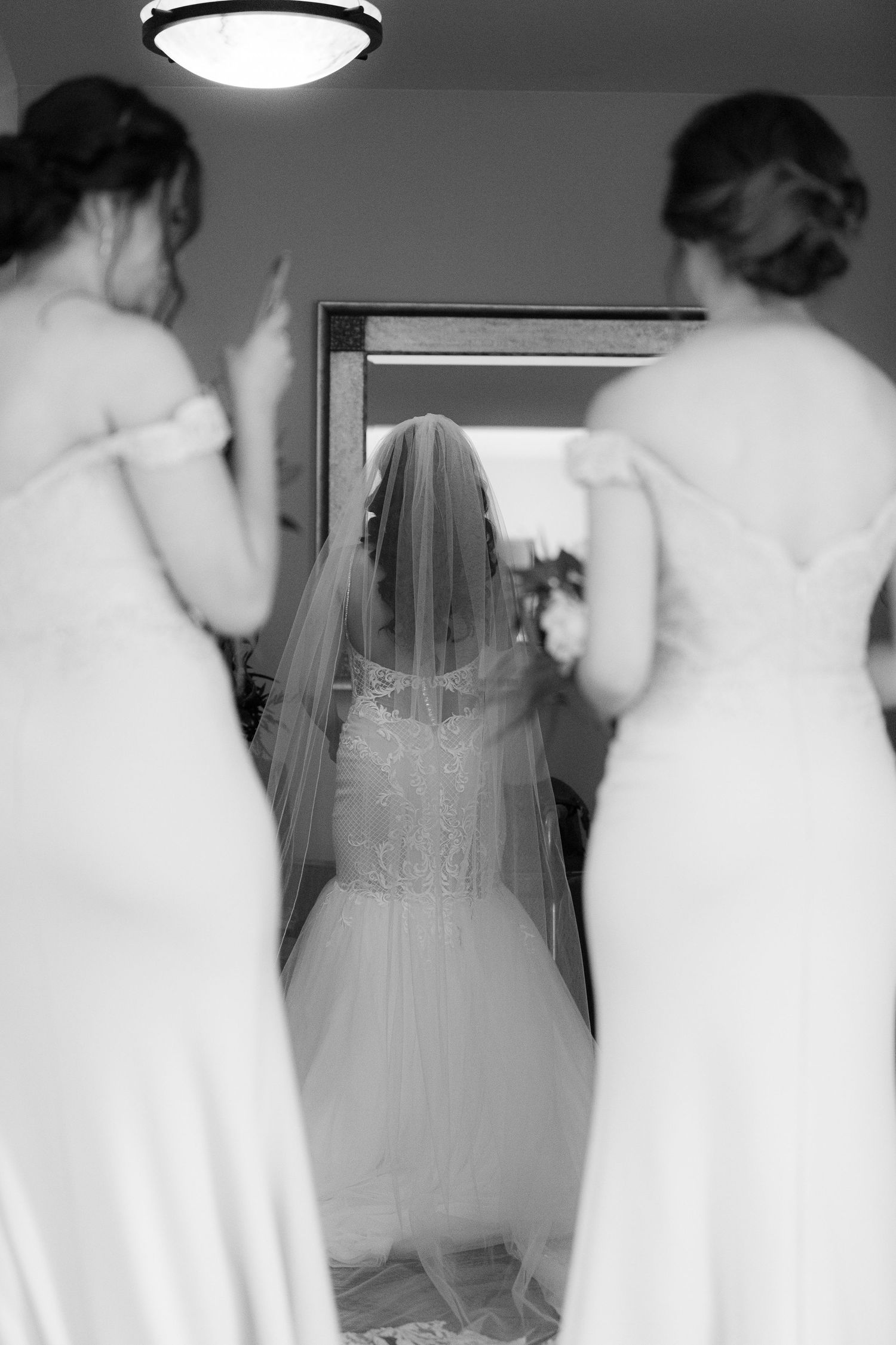 Several women in white dresses preparing in a bridal suite with a full-length mirror.