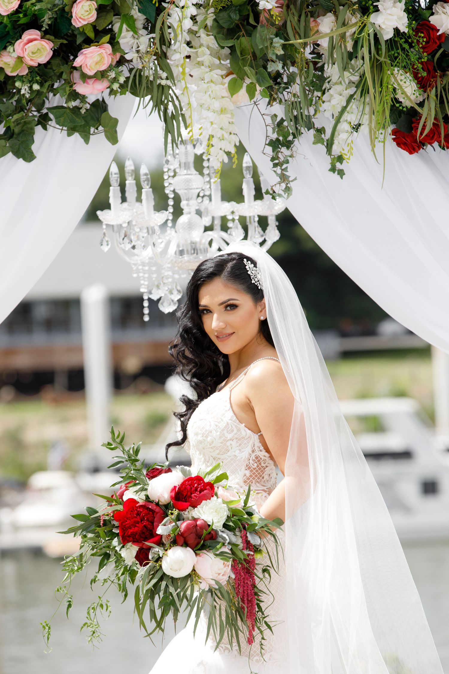 Elegant bridal bouquet with red and white flowers under crystal chandelier and floral arch with draped white fabric.