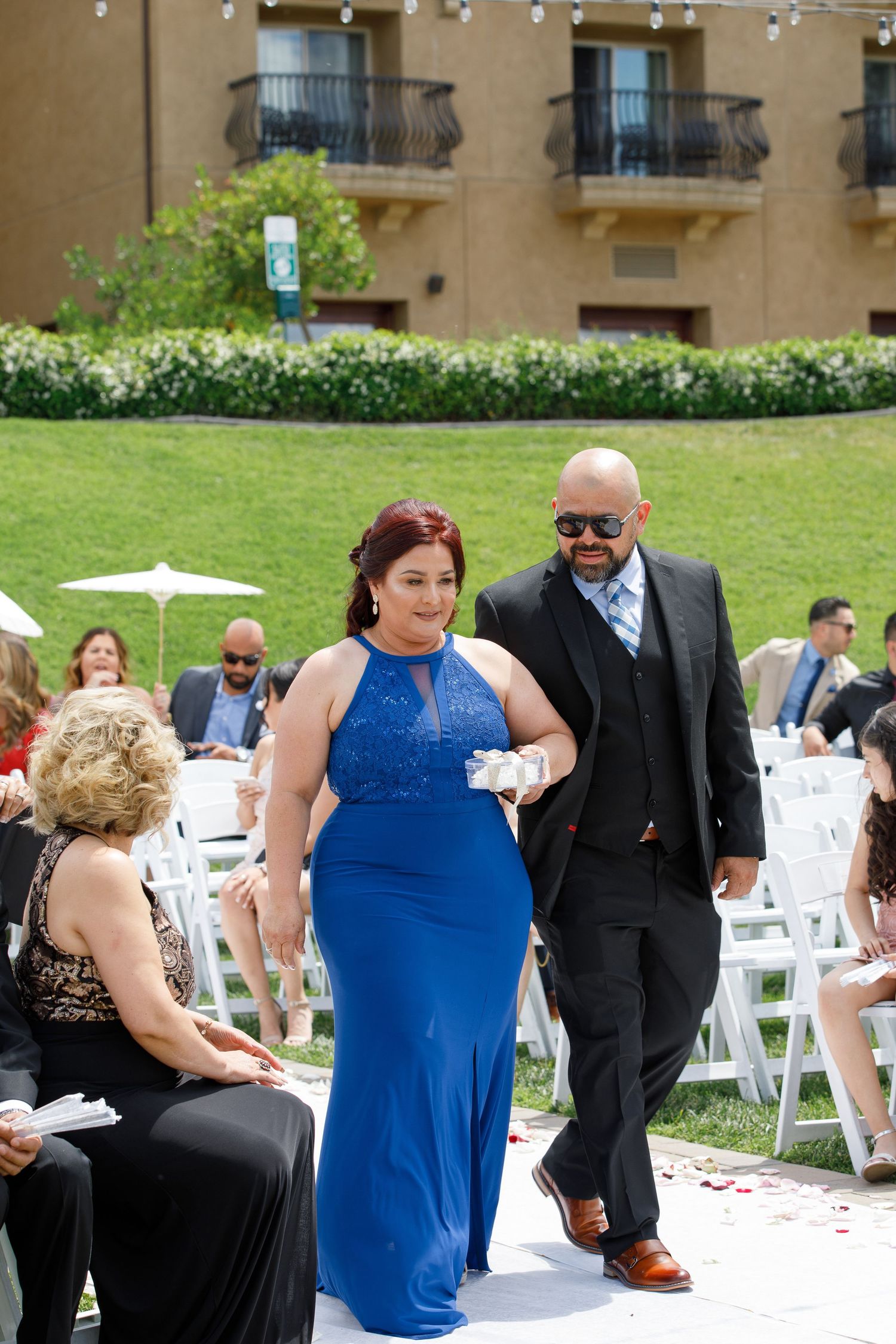 Wedding guests in formal attire mingle on a lawn with white chairs set up for an outdoor ceremony.