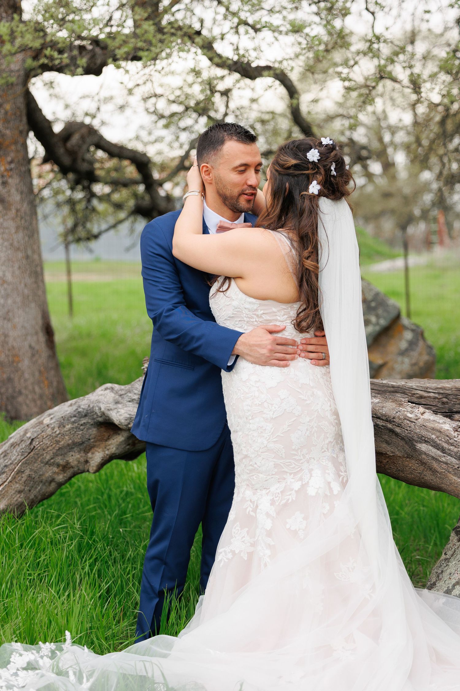 A romantic wedding couple embraces under blooming spring trees in an outdoor garden setting.