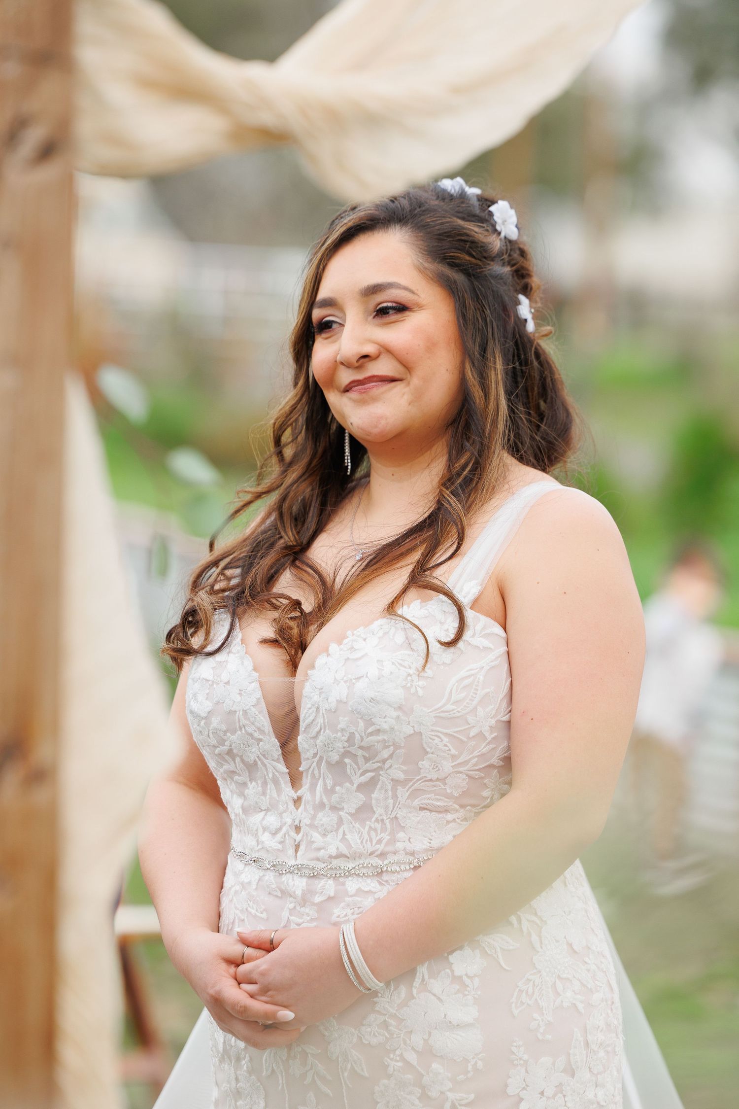 A bride in a white lace wedding dress poses outdoors with natural lighting and greenery in the background.