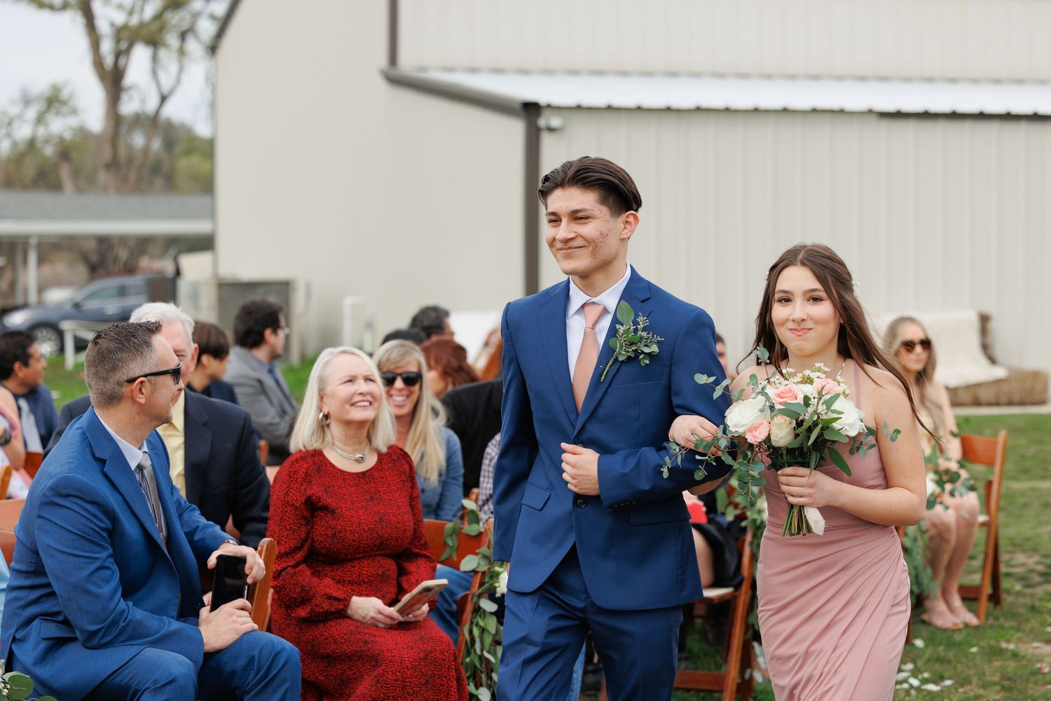 A wedding party poses in formal attire with navy suits and burgundy dresses at an outdoor venue.
