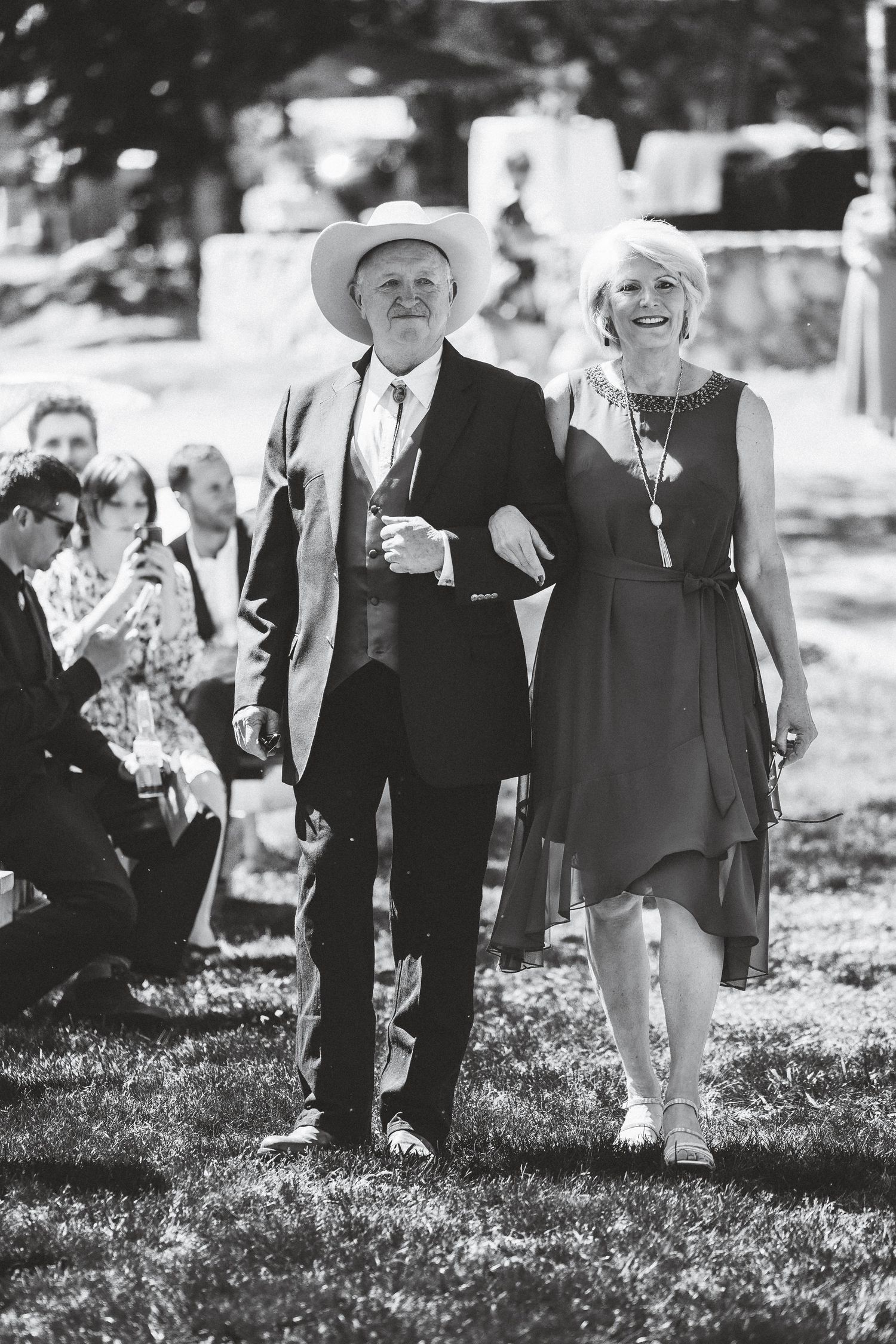 Wedding guests walk through a grassy lawn during an outdoor celebration with sunlight filtering through the trees.