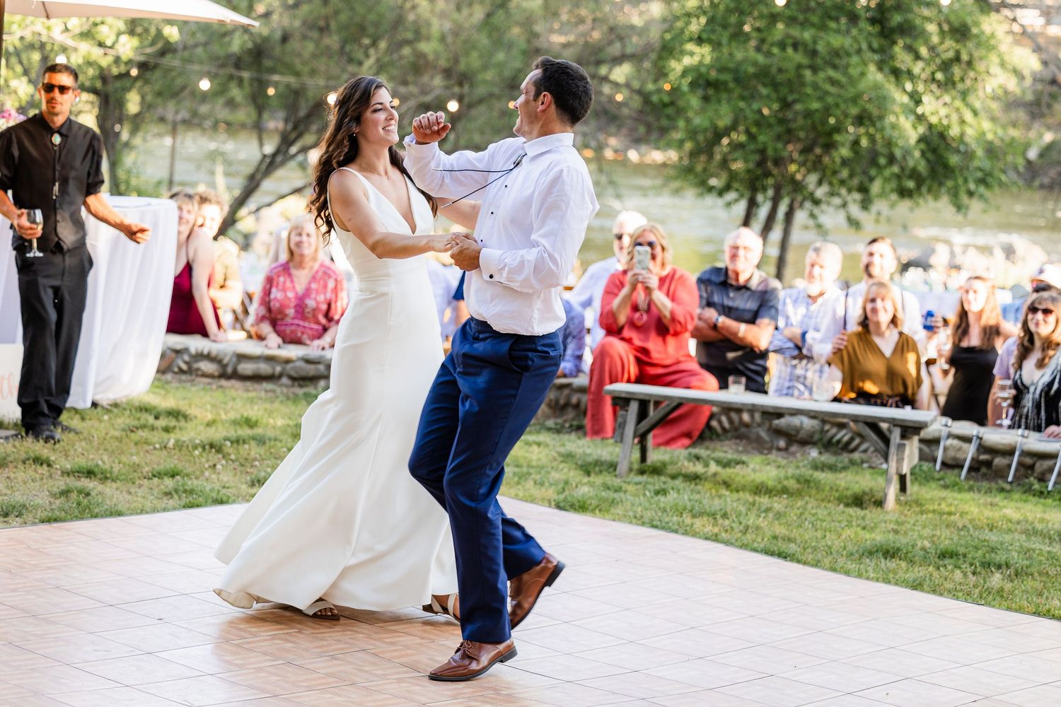 A couple dances together on a sunny outdoor patio during a celebration with guests watching in the background.