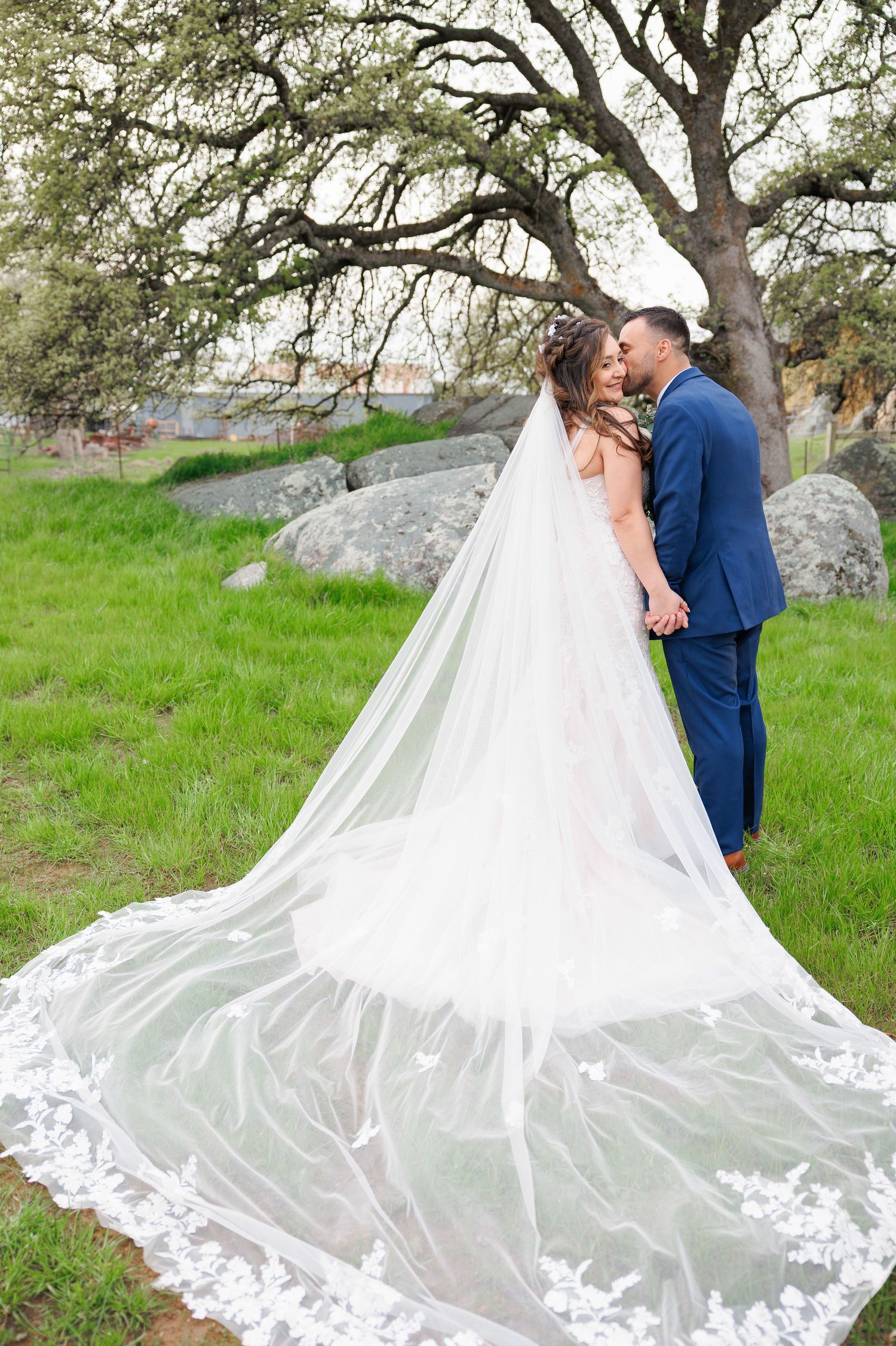 A romantic wedding couple embraces on a grassy hillside as a long white veil flows dramatically behind them.