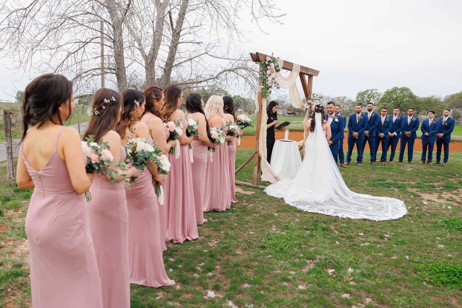 Wedding party in pink bridesmaid dresses and navy suits stands together for outdoor photos on a spring day.