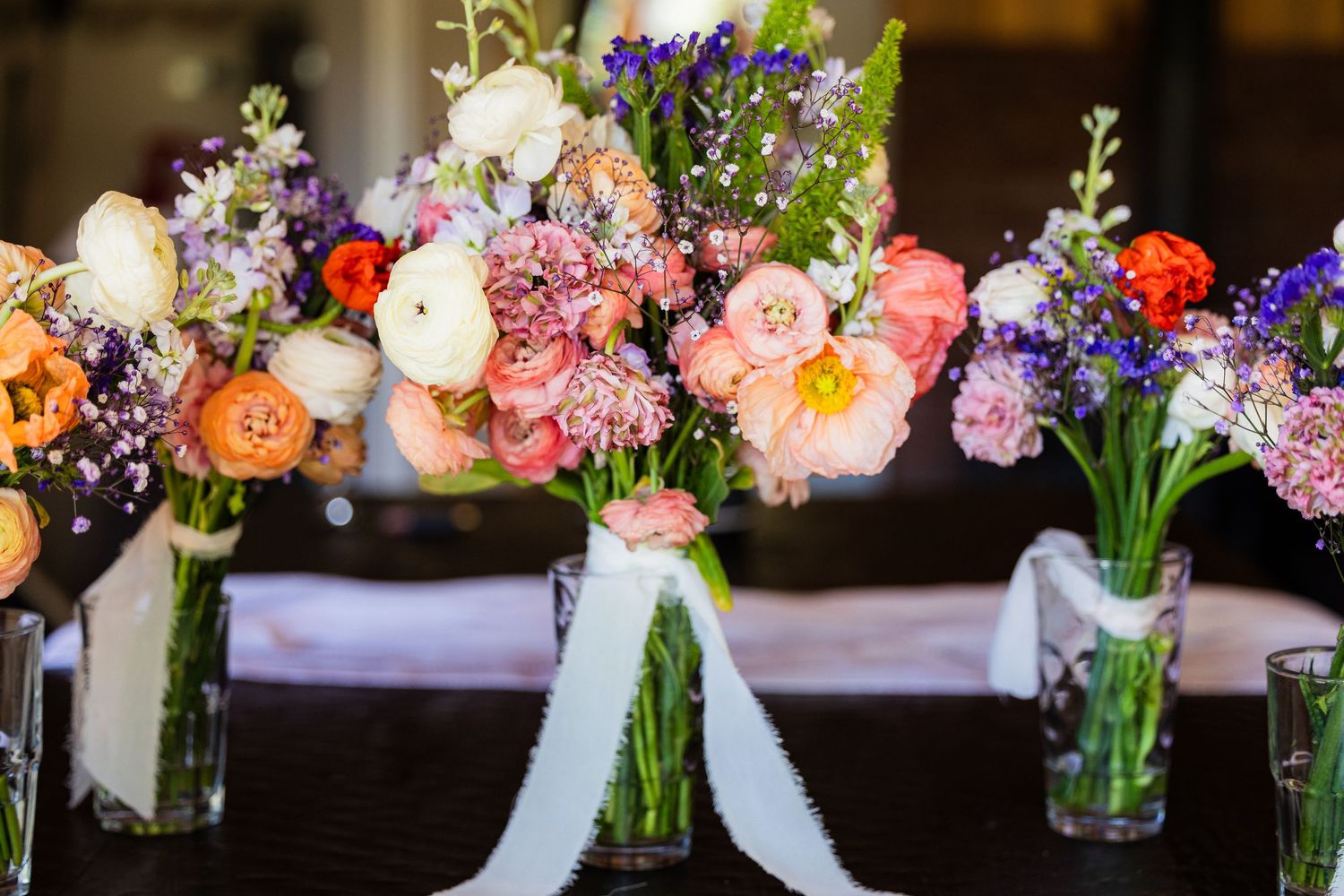 Mason jars filled with colorful spring flowers line a rustic wooden table at a wedding reception.
