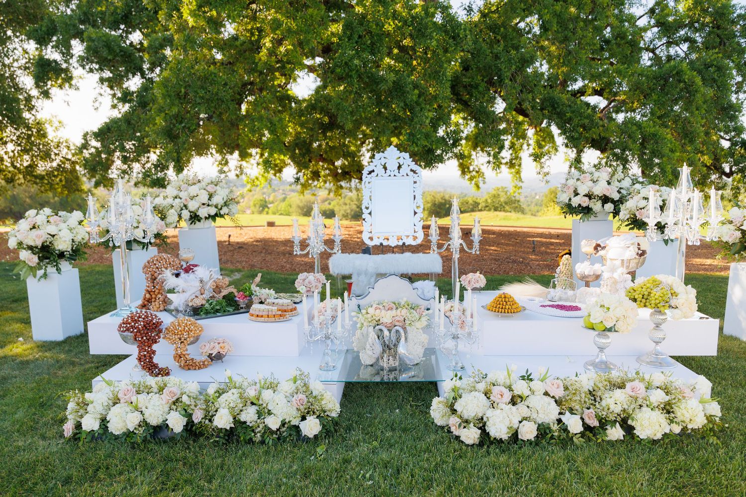 Elegant outdoor wedding reception setup with white linens, floral centerpieces and string lights under trees at sunset.