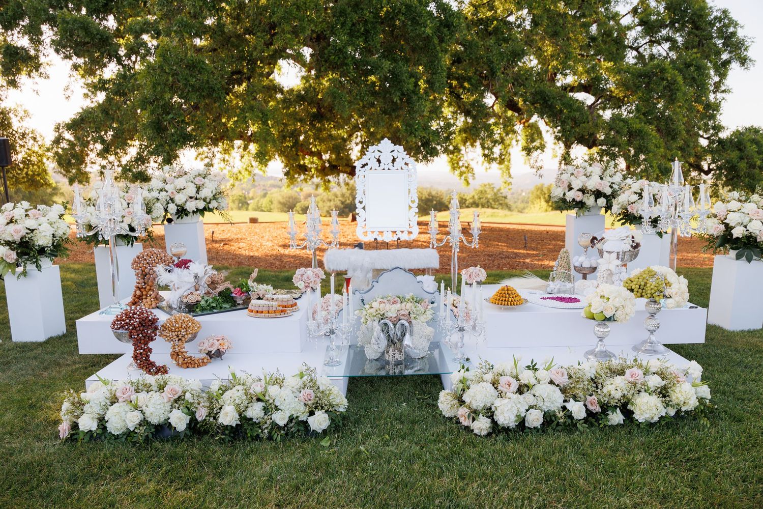 Elegant outdoor wedding reception setup with white floral arrangements and glowing candles at sunset.