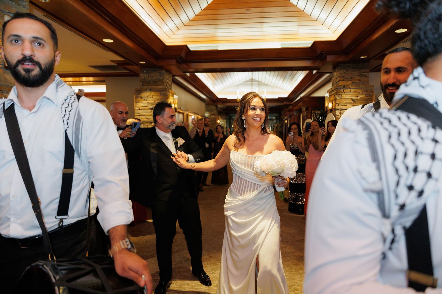 Wedding guests dance and celebrate together at an indoor reception with wooden ceiling beams.