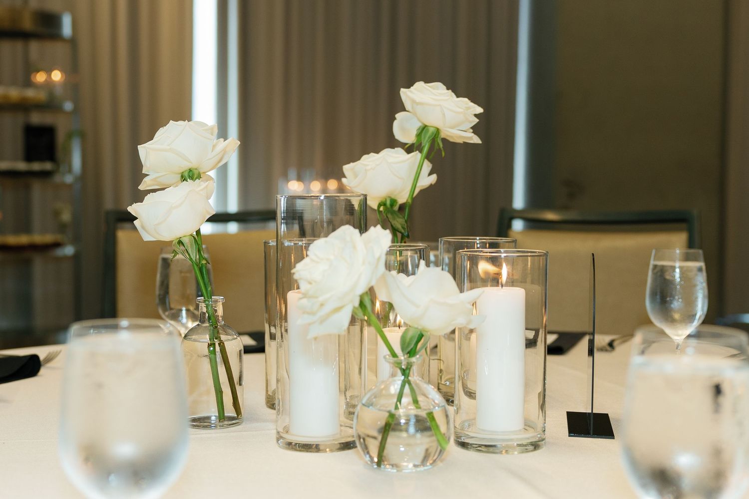 White rose centerpieces in glass vases adorn tables with crisp white linens and black accents at a formal dining setup.