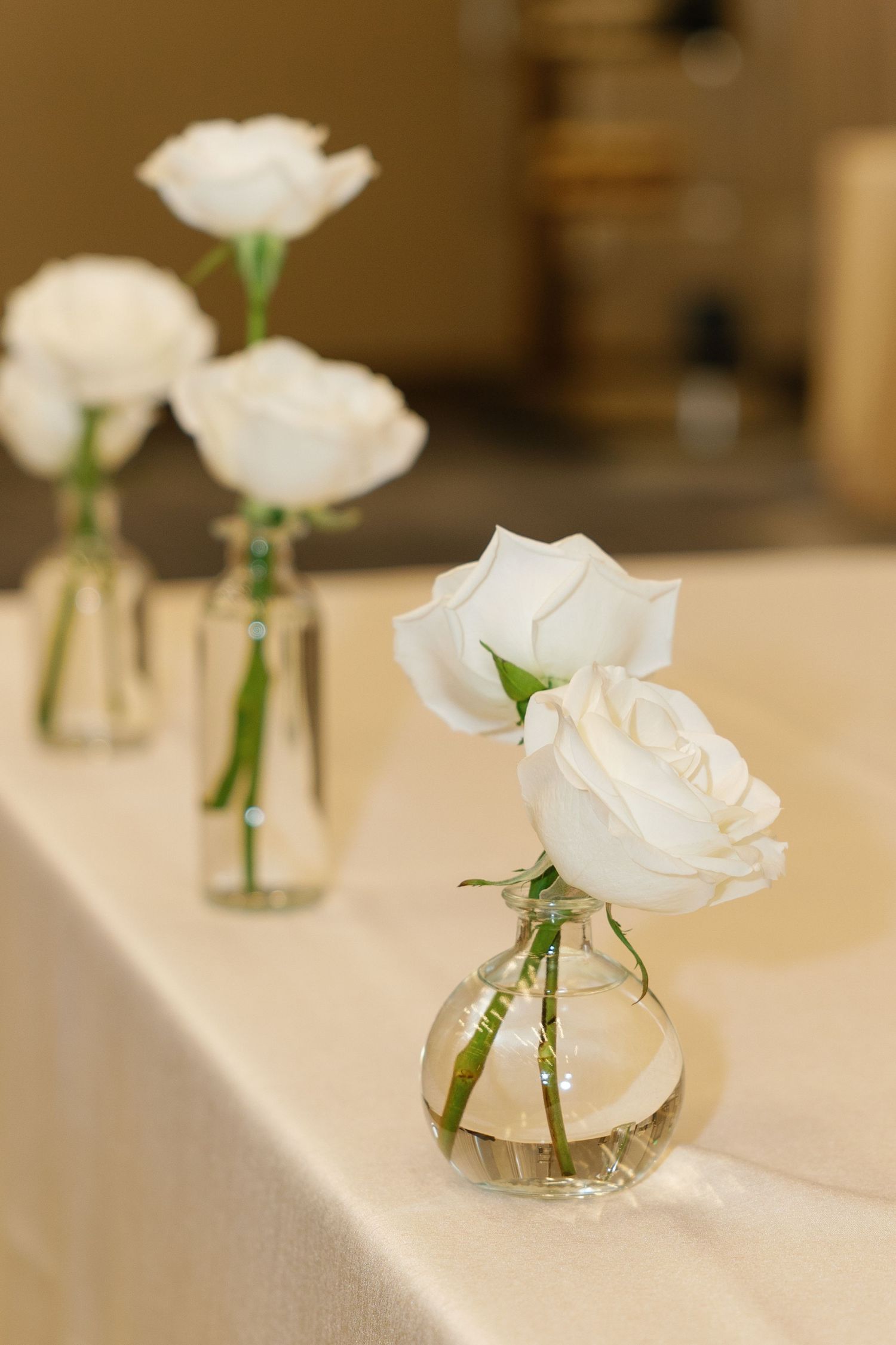 White roses in small glass vases arranged on a white tablecloth create an elegant wedding table setting.
