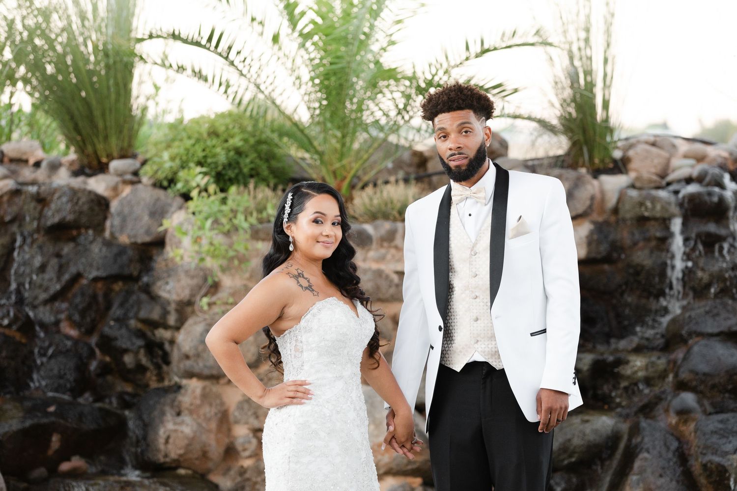 A couple in wedding attire poses together against a natural stone backdrop with lush greenery.