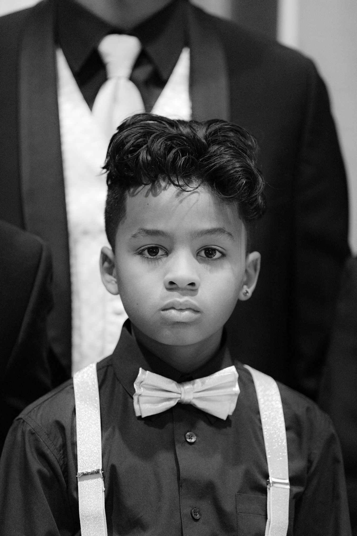 Black and white portrait of a young child wearing a dark shirt with white suspenders and a bow tie.