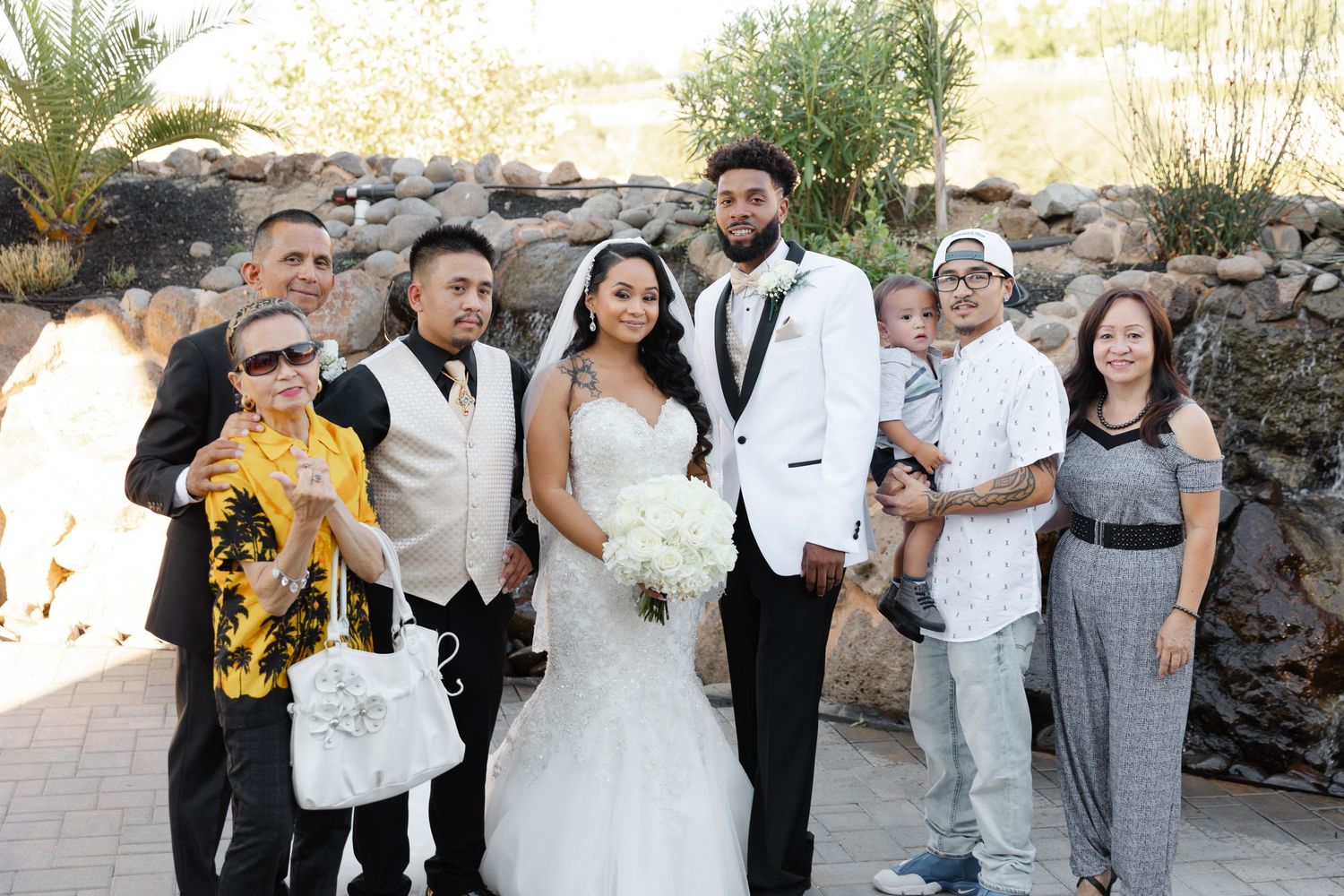 Wedding party members pose together outdoors in formal attire for group photos near a stone wall.