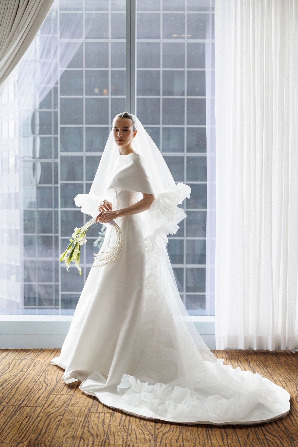 A bride in a classic white wedding gown poses elegantly by sunlit windows in a bright room.
