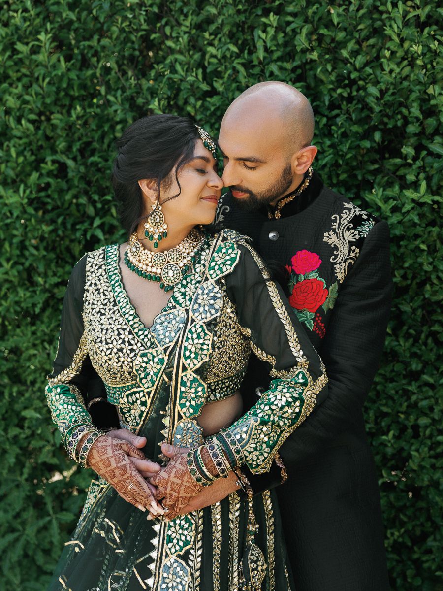 A couple in traditional South Asian wedding attire embraces against a lush green hedge backdrop.